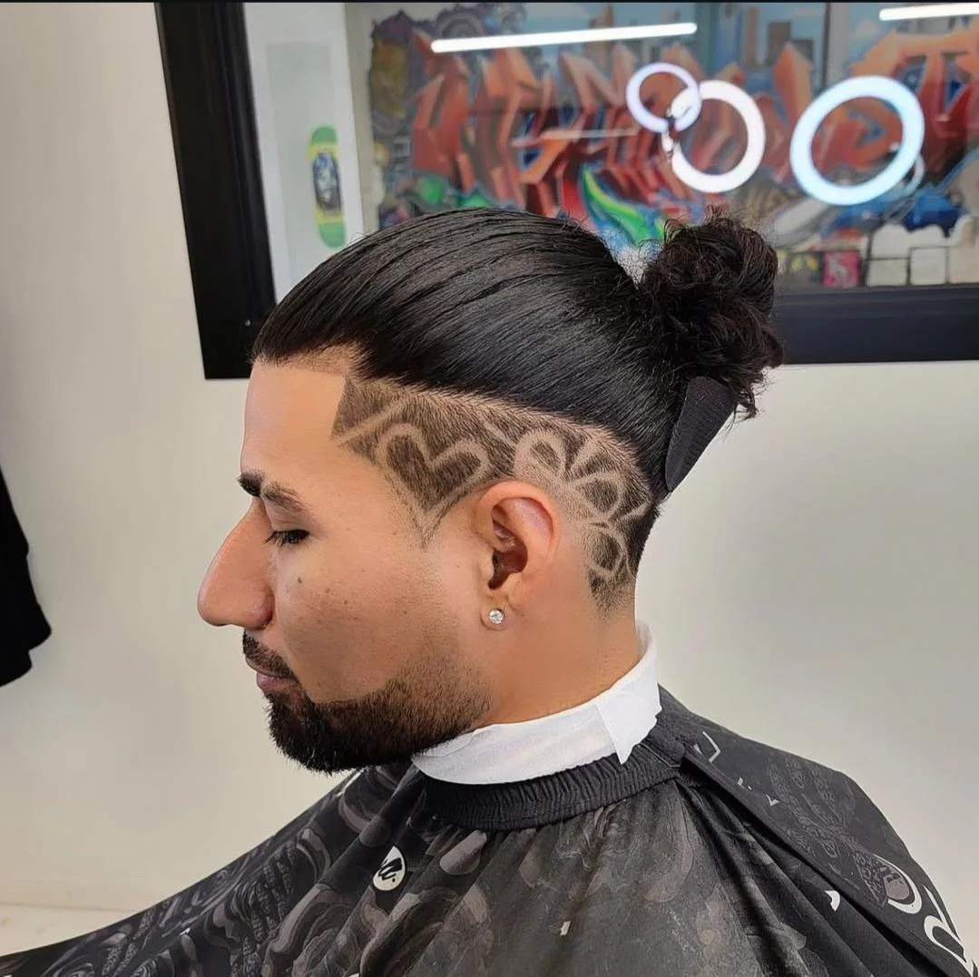 Man with a slicked-back hairstyle featuring shaved geometric designs on the side, sitting in a barber's chair in a salon.