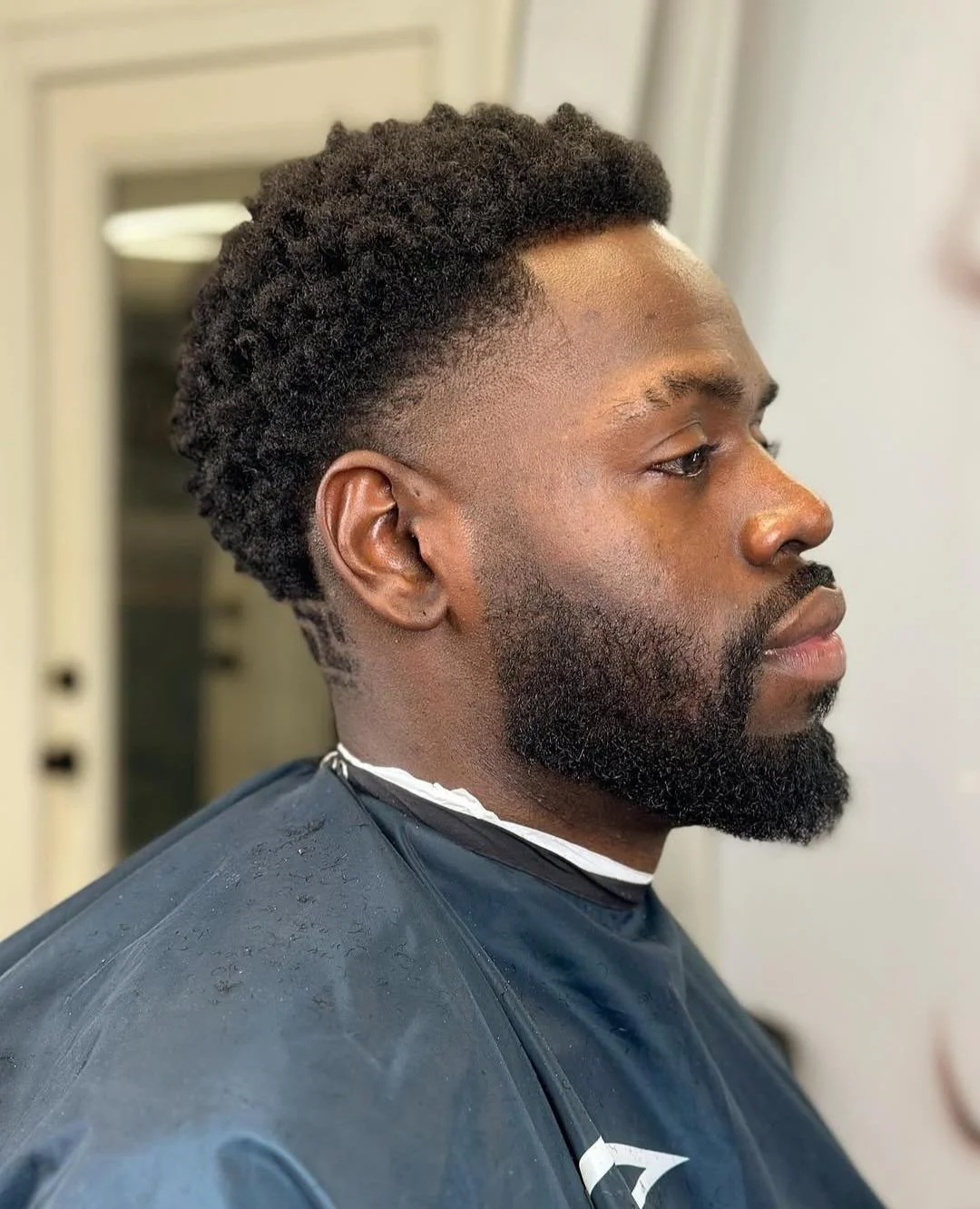 Side profile of a man with a textured, curly hairstyle, a full beard, and a fade haircut, sitting in a barber chair draped with a black cape.