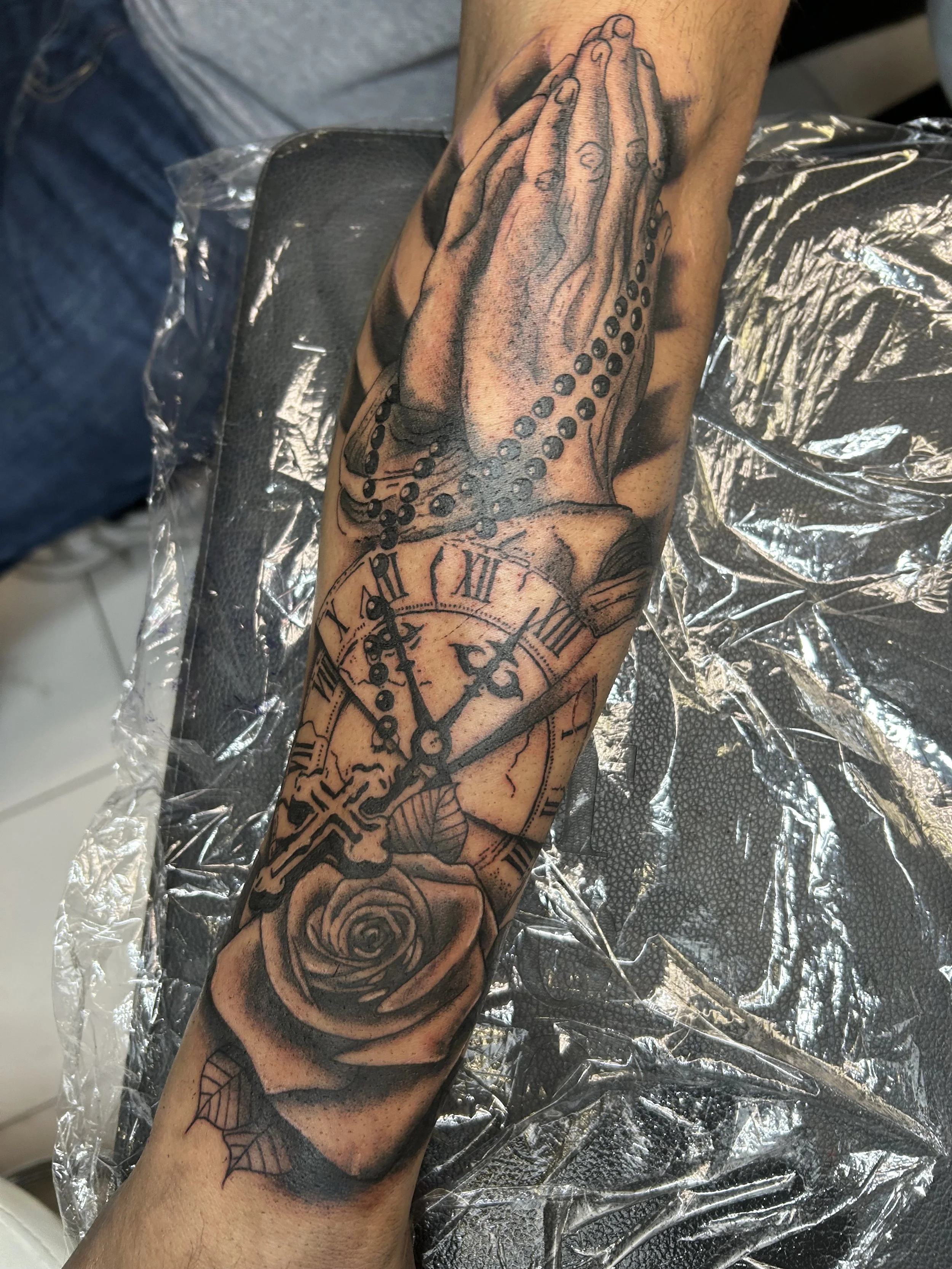 Tattoo of praying hands holding rosary, a clock showing 1:20, and a rose.