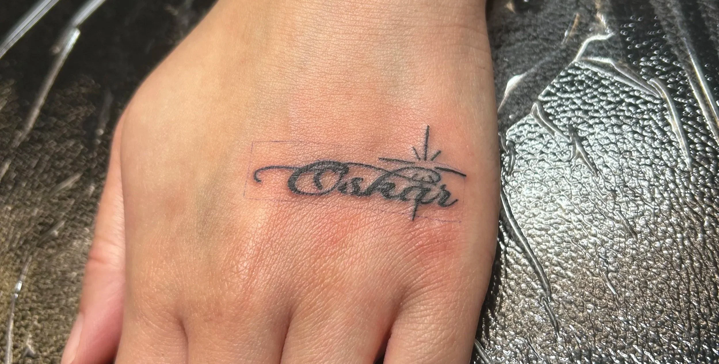 Tattoo of the word 'family' in cursive script on a person's hand, with a star-like design above the 'i'.