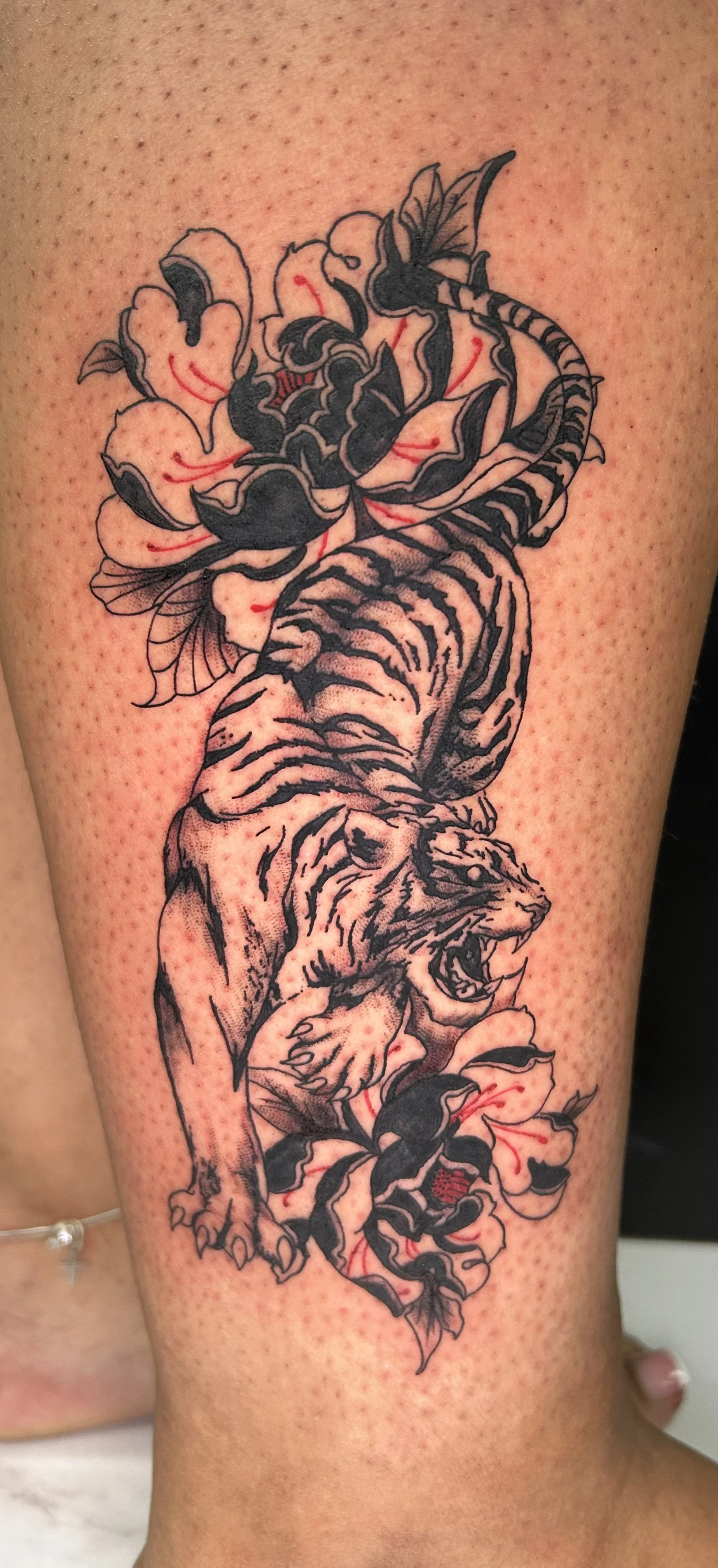 A tattoo of a roaring lion with a tiger's body, surrounded by flowers and leaves, inked in black and gray shading on a person's arm.