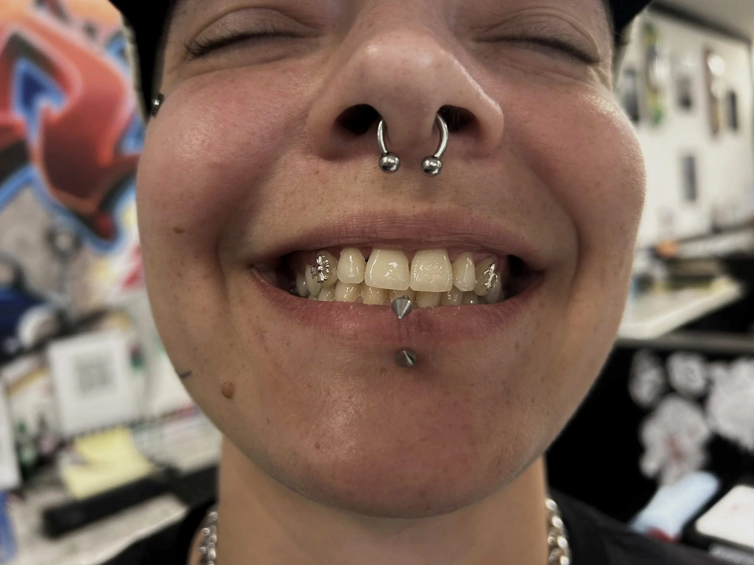 Close-up of a person with multiple facial piercings, including a septum ring, two lower lip piercings, and a ring on the nose. They have braces on their teeth and are smiling with eyes closed.