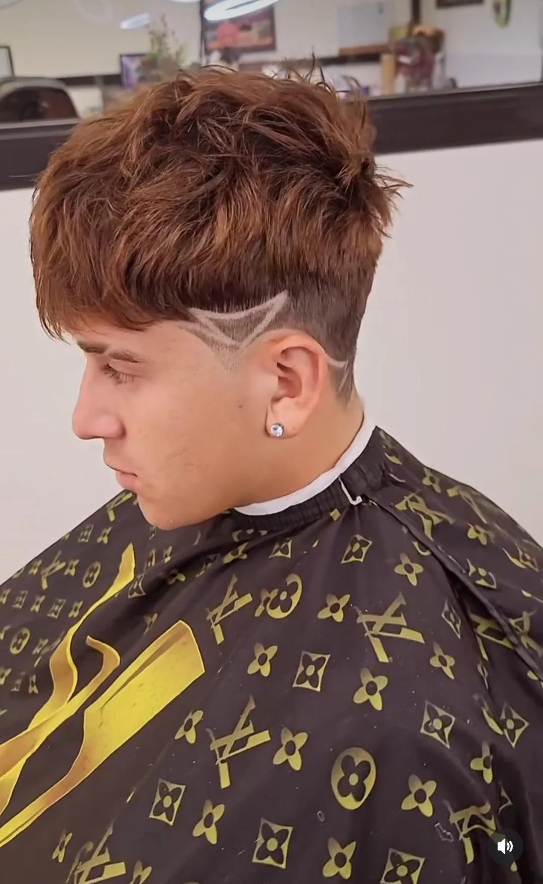 A young man with styled auburn hair sitting in a barber's chair, wearing a patterned cape, with a design shaved into the side of his head.