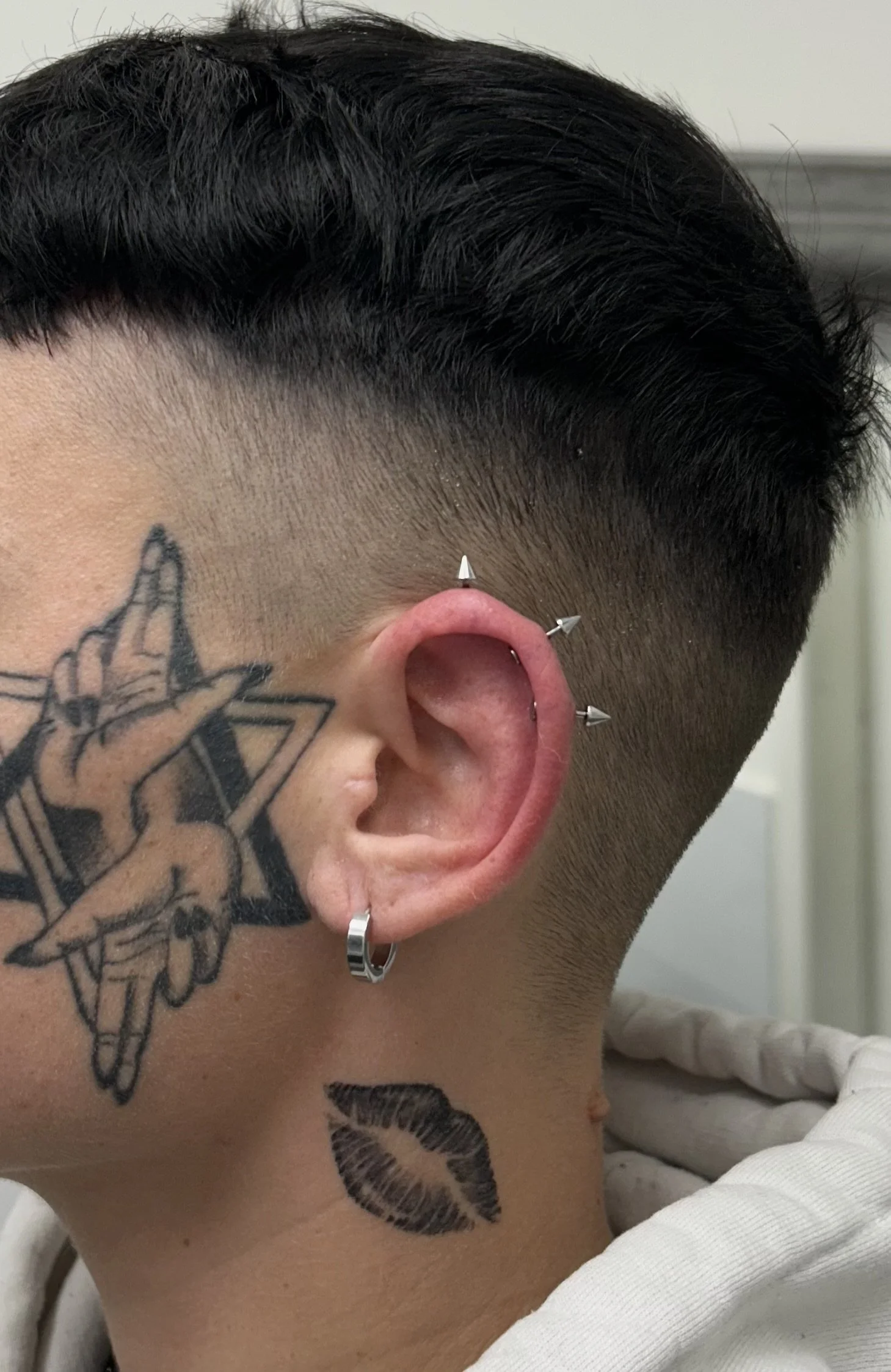Close-up of a person's ear with multiple piercings, including an ear cuff, and tattoos on their neck and face, with a short, dark hairstyle.