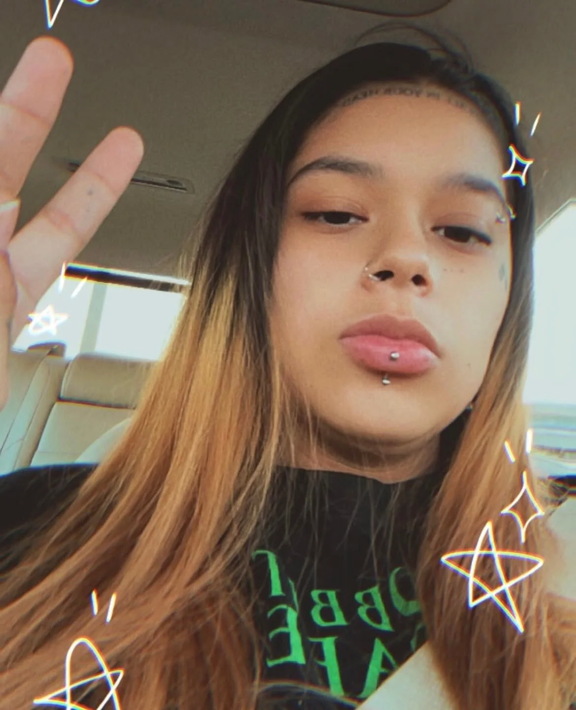 A young woman with long ombré hair making a peace sign gesture. She has a nose ring, a lip piercing, and a facial tattoo near her eye. She is wearing a black shirt with green text. The background shows the interior of a vehicle, with beige seats and windows, and a sunny outdoor scene.