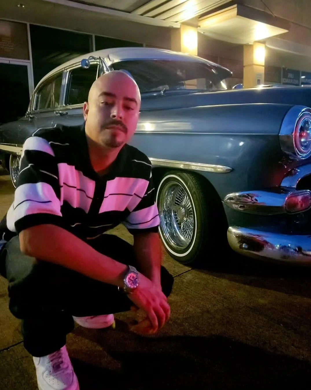 A man crouching in front of a vintage black car at night.