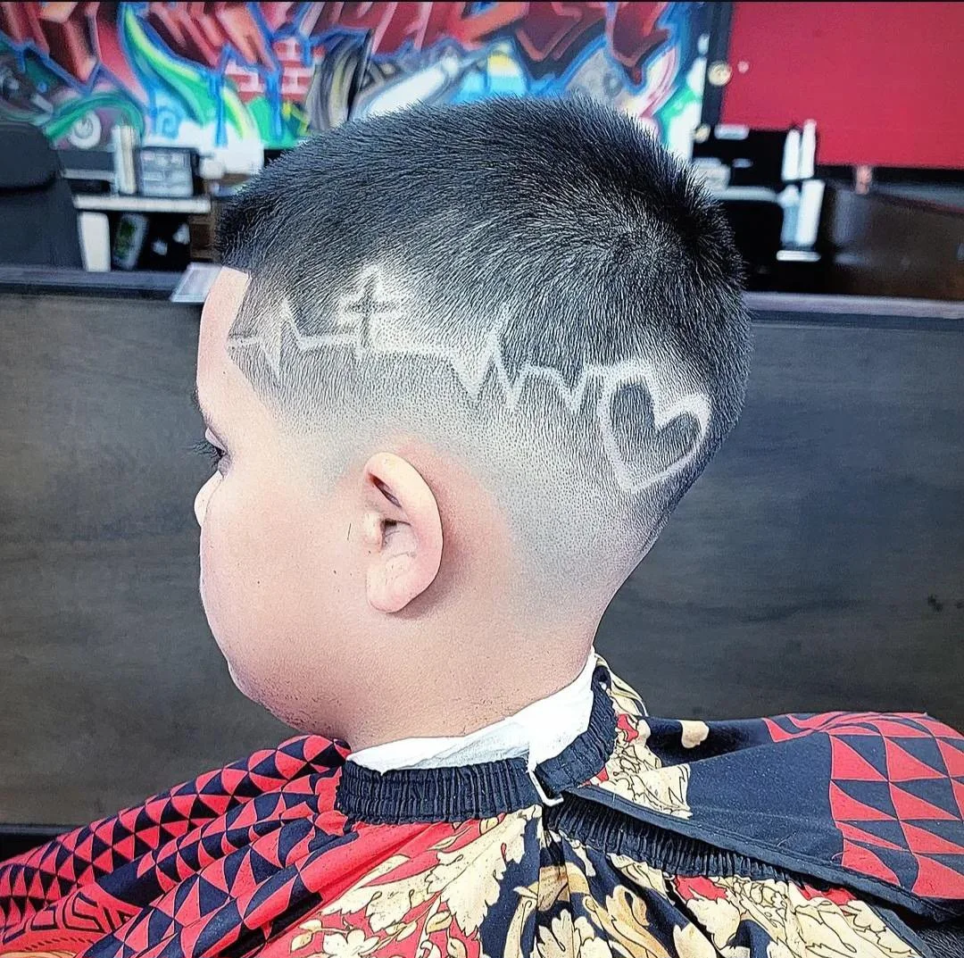 A young boy with a modern buzz haircut featuring a shaved design of a heart and an electrocardiogram line etched into the side of his head, sitting in a barber's chair with a patterned cape, in front of a colorful graffiti wall.