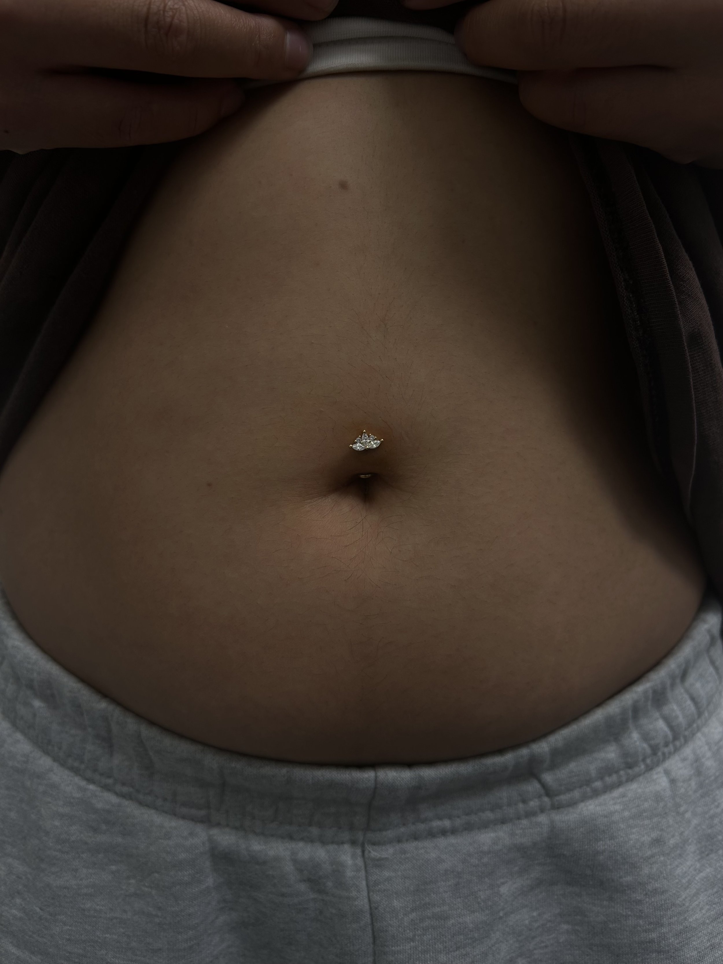 Close-up of a person's lower abdomen showing a navel piercing with a shiny, floral-shaped gemstone jewelry, wearing gray clothing.