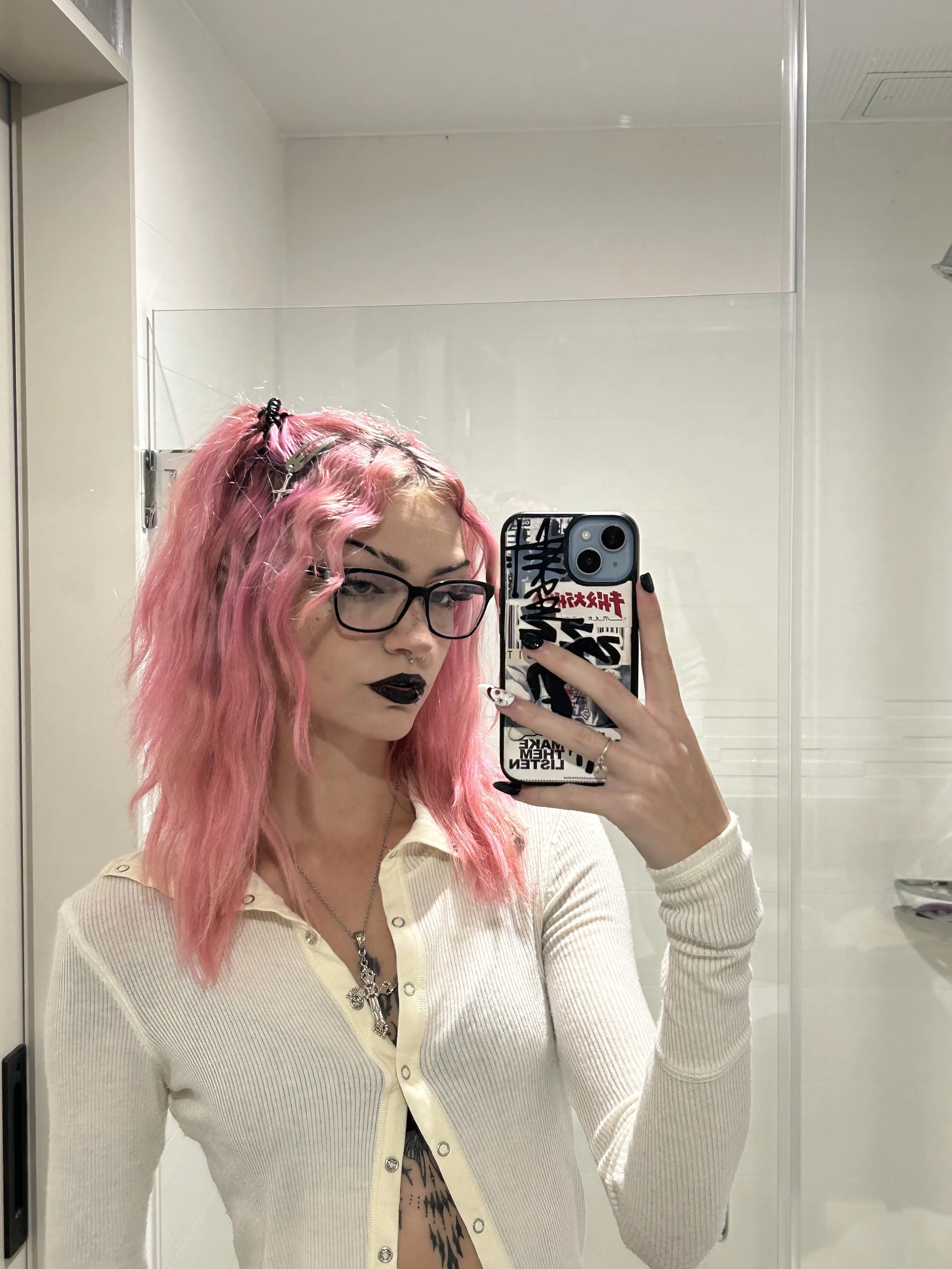 Young woman with pink curly hair, black lipstick, glasses, and jewelry taking a selfie in a bathroom mirror.
