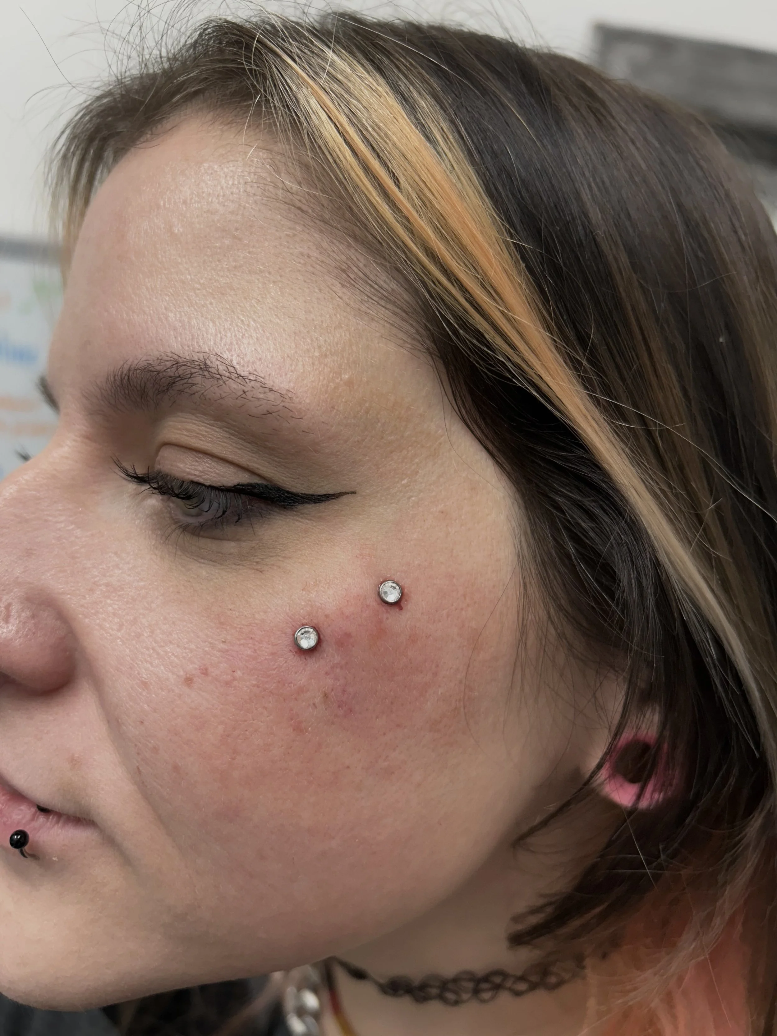 Close-up of a woman's face showing facial piercings, including two dermal piercings with gemstone ends on her cheek, a black lip piercing, and a pink ear piercing, with makeup including winged eyeliner and subtle eye shadow.
