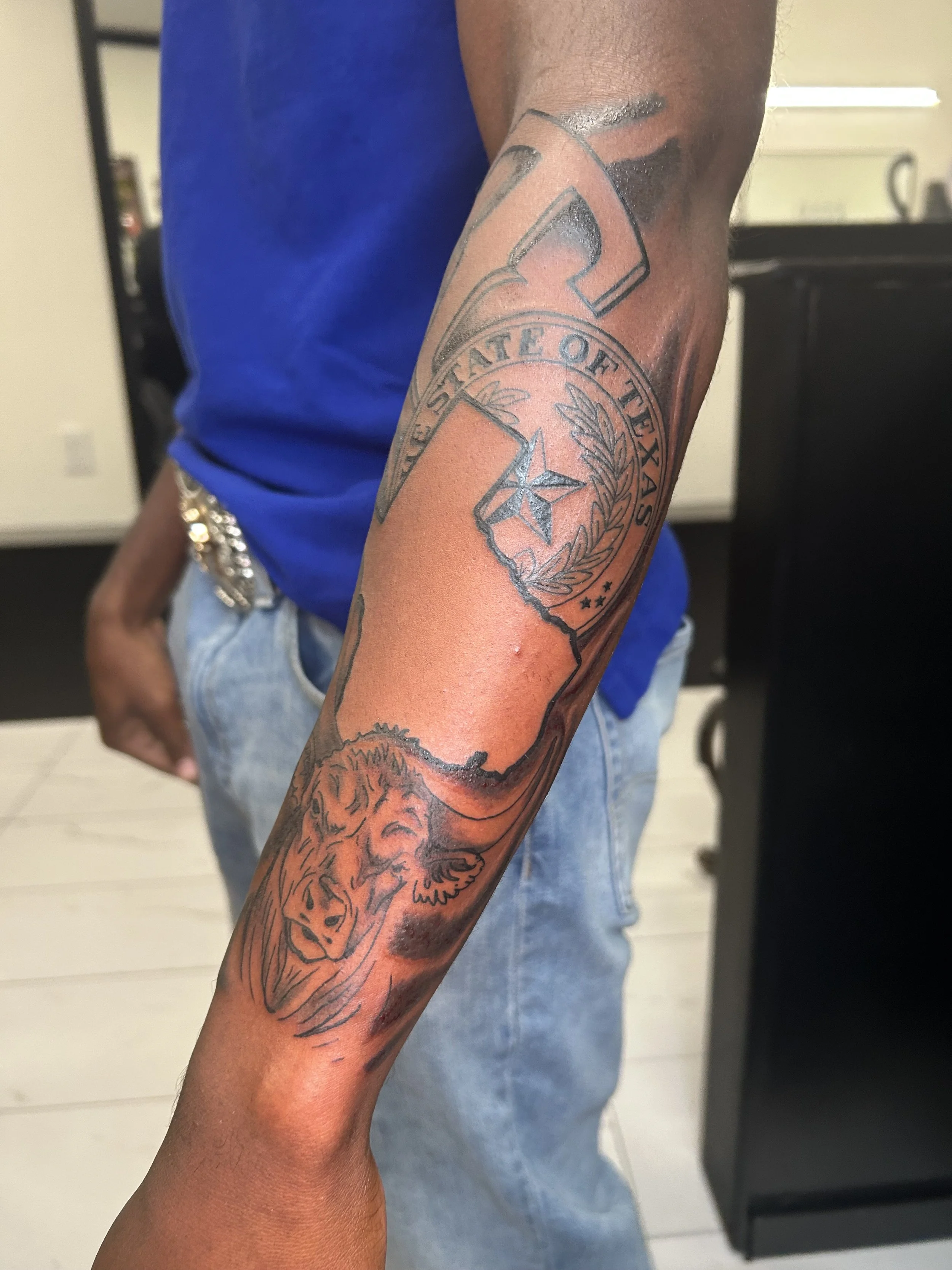 Tattoo on person's arm depicting the state of Texas with a star, a wreath, and the phrase 'State of Texas', alongside a lion's head.