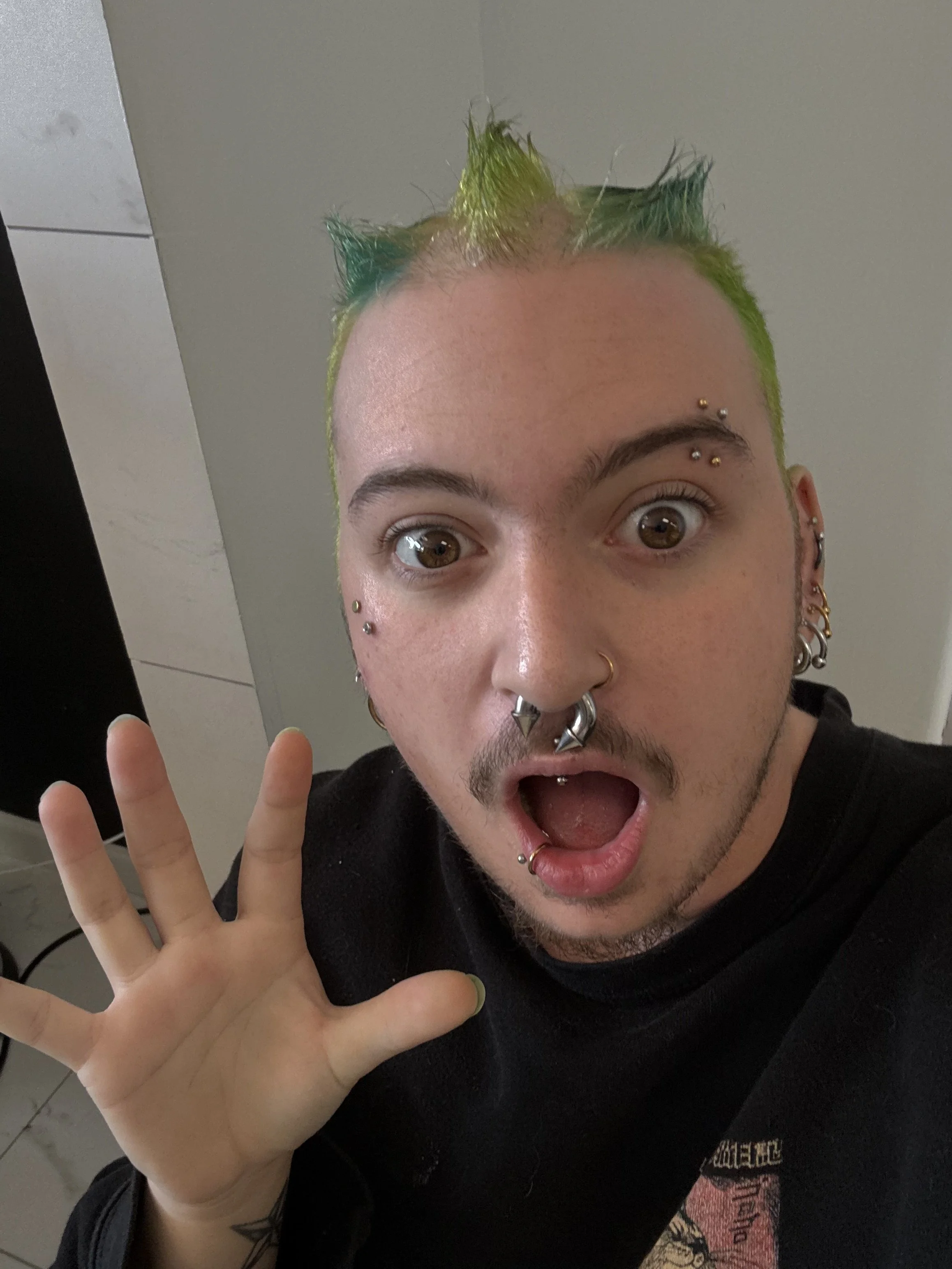 Young person with vibrant green and blue hair, multiple facial piercings, and surprised expression, holding up a hand in front of camera.