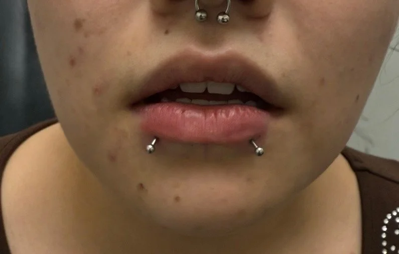 Close-up of a person with facial piercings on the lower lip and a nose ring, showing part of the mouth and chin.