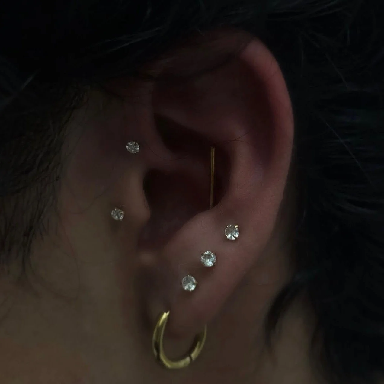 Close-up of a person's ear with multiple earrings, including small diamond studs and gold hoops, and a gold straight bar earring.