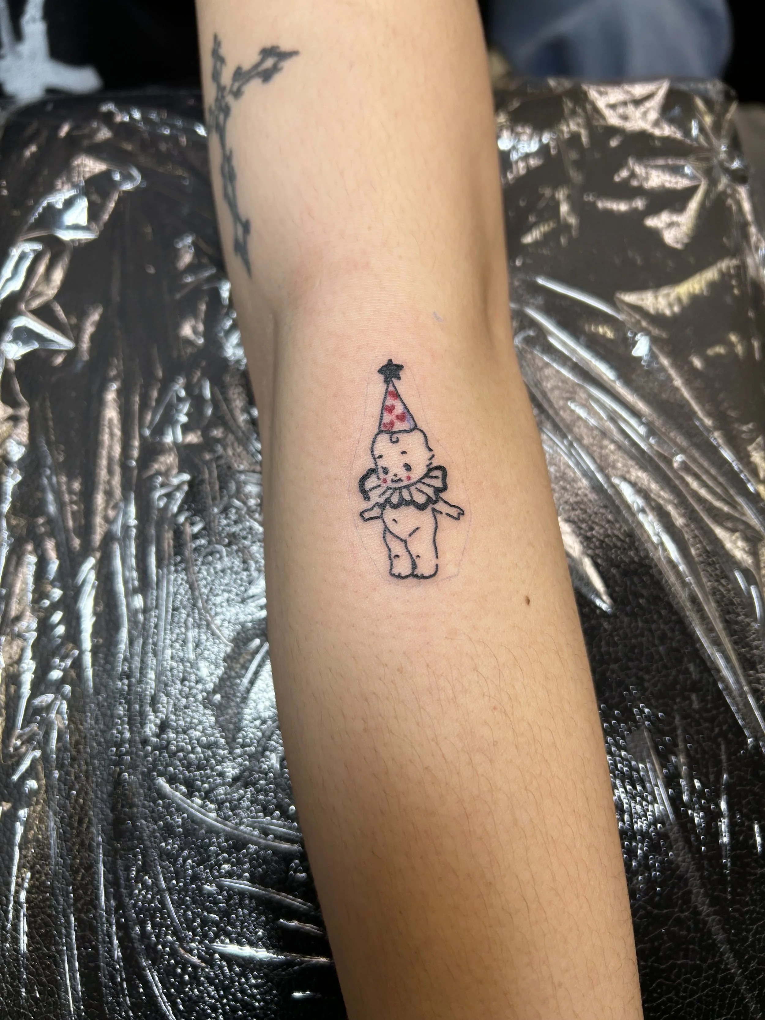 Small tattoo of a cute elephant wearing a ruffled collar and a party hat with a star top on a person's forearm.