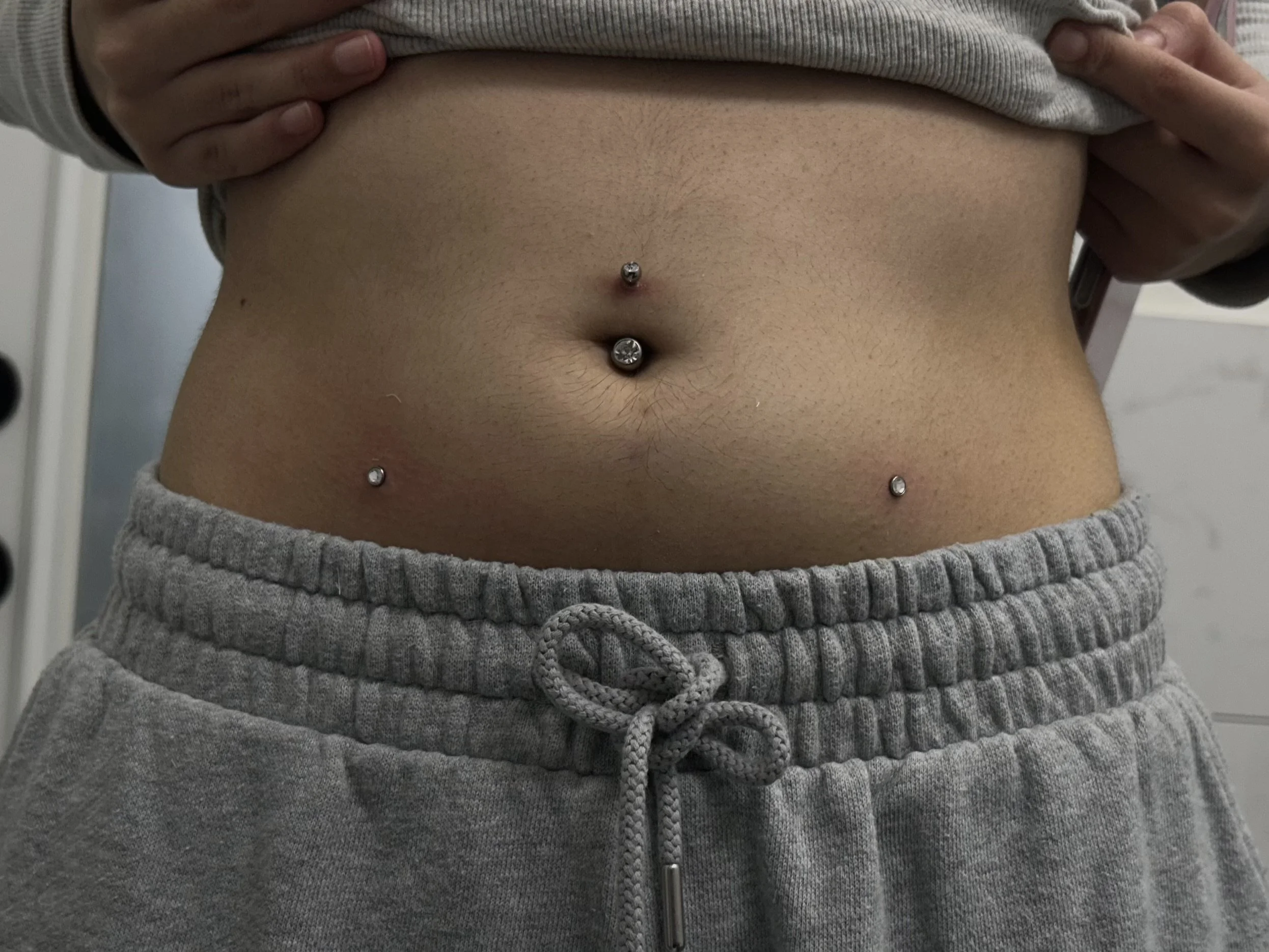 A person's midsection showing a navel piercing with three jewelry pieces and gray sweatpants with a drawstring, lifting a gray shirt to reveal the abdomen.