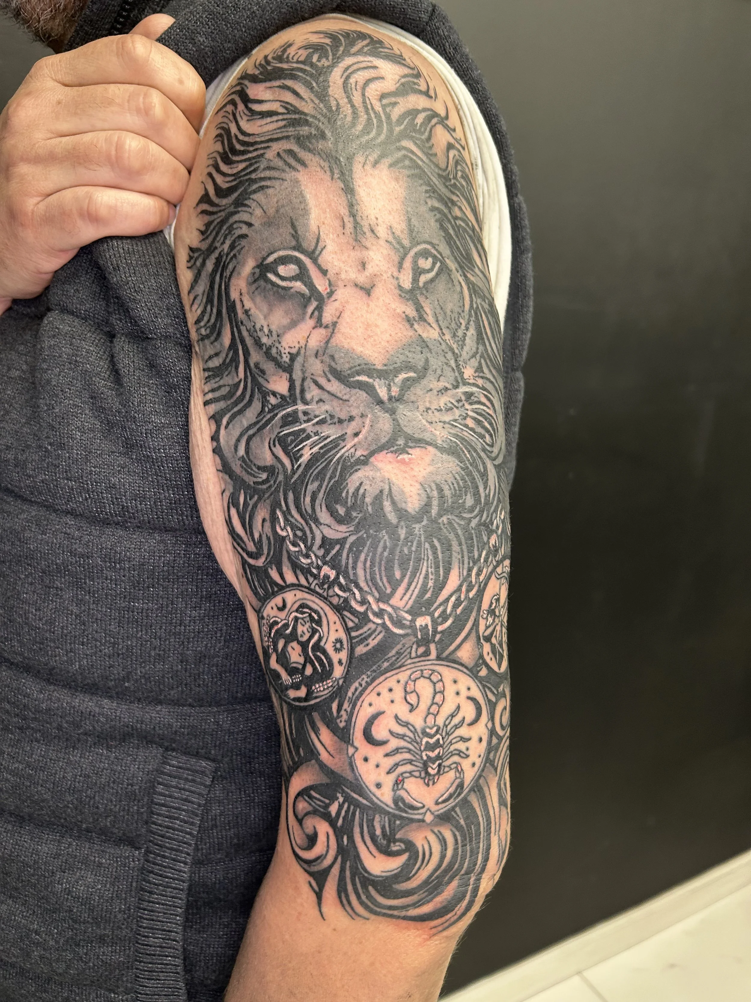 Tattoo of a lion's face on a person's upper arm, in black and gray ink, with some additional smaller tattoo designs below.
