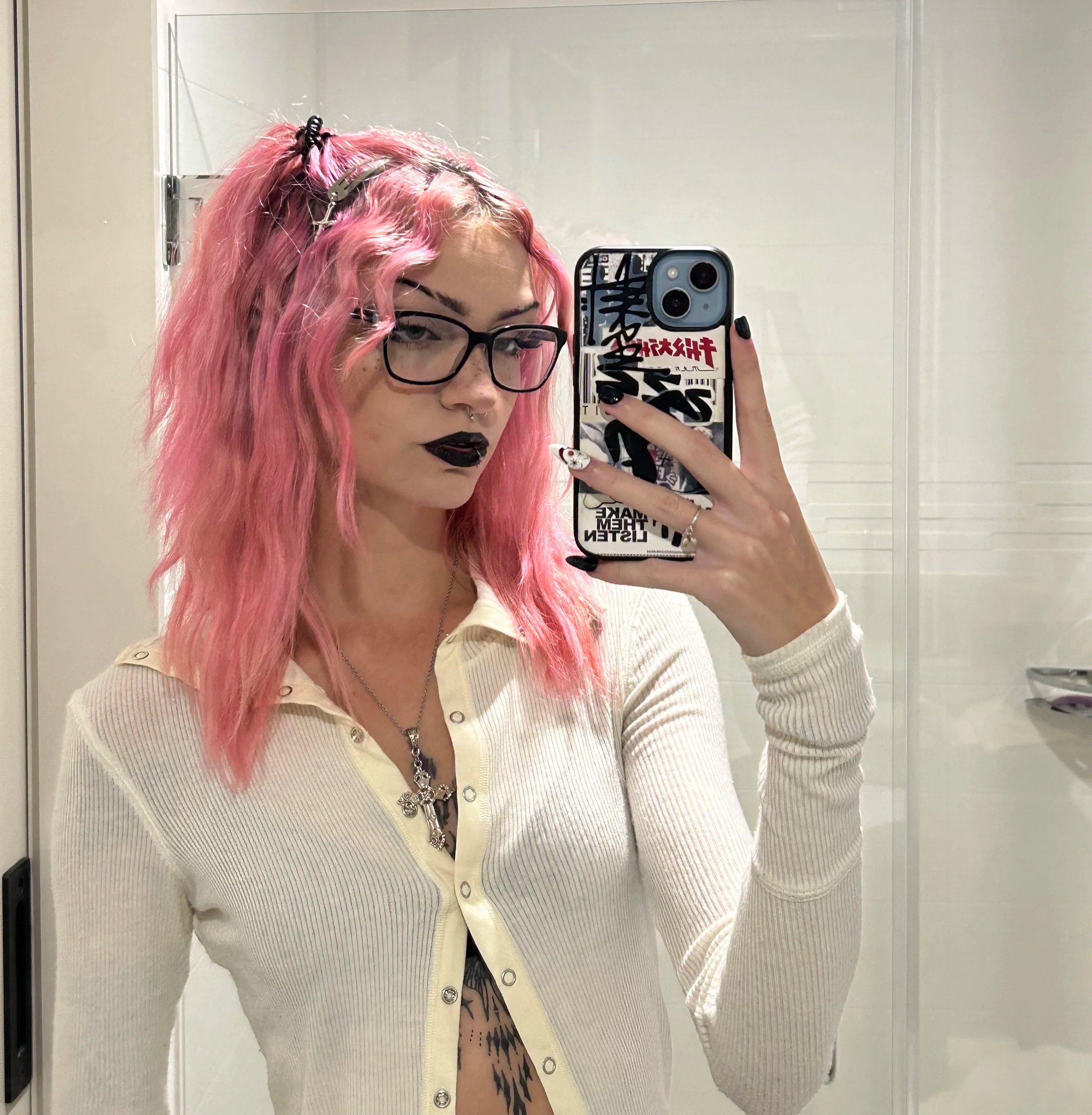woman with pink wavy hair, glasses, black lipstick, and piercings taking a mirror selfie in a bathroom