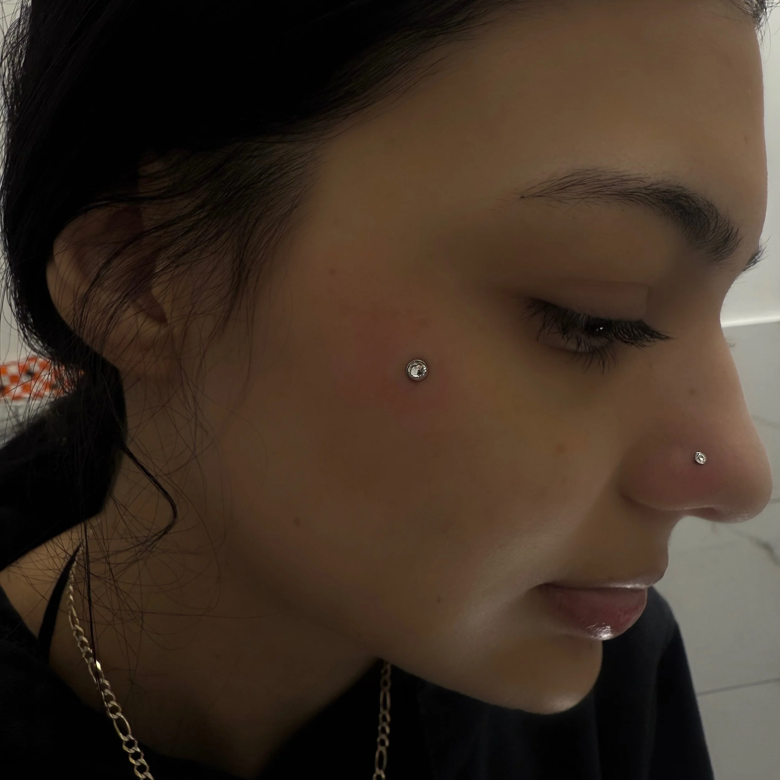 Close-up of a woman's face in profile, with two nose piercings and face jewelry, with dark hair and light skin.