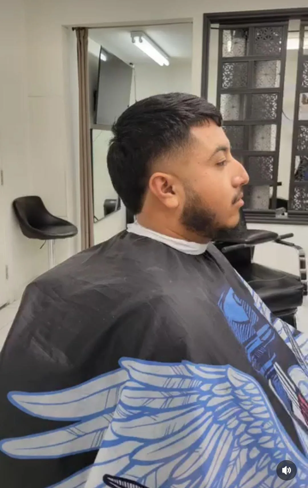 A man with dark hair and a beard sitting in a hair salon chair, wearing a black cape with a blue and white design, in front of a mirror.