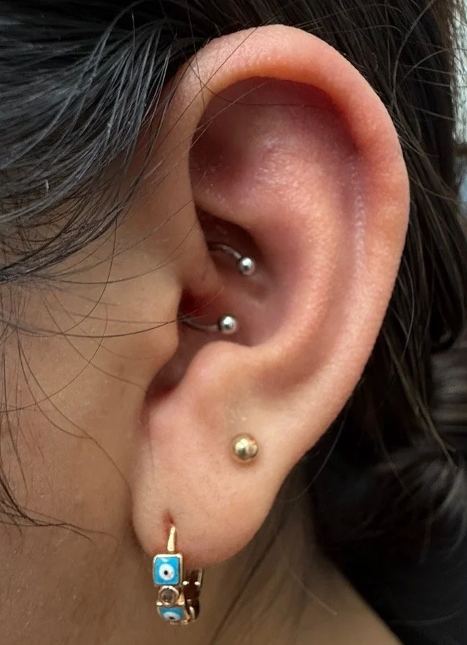 Close-up of a person's ear with multiple piercings, including a blue evil eye charm earring, two silver studs, and a gold ball stud, with dark hair partially covering the ear.