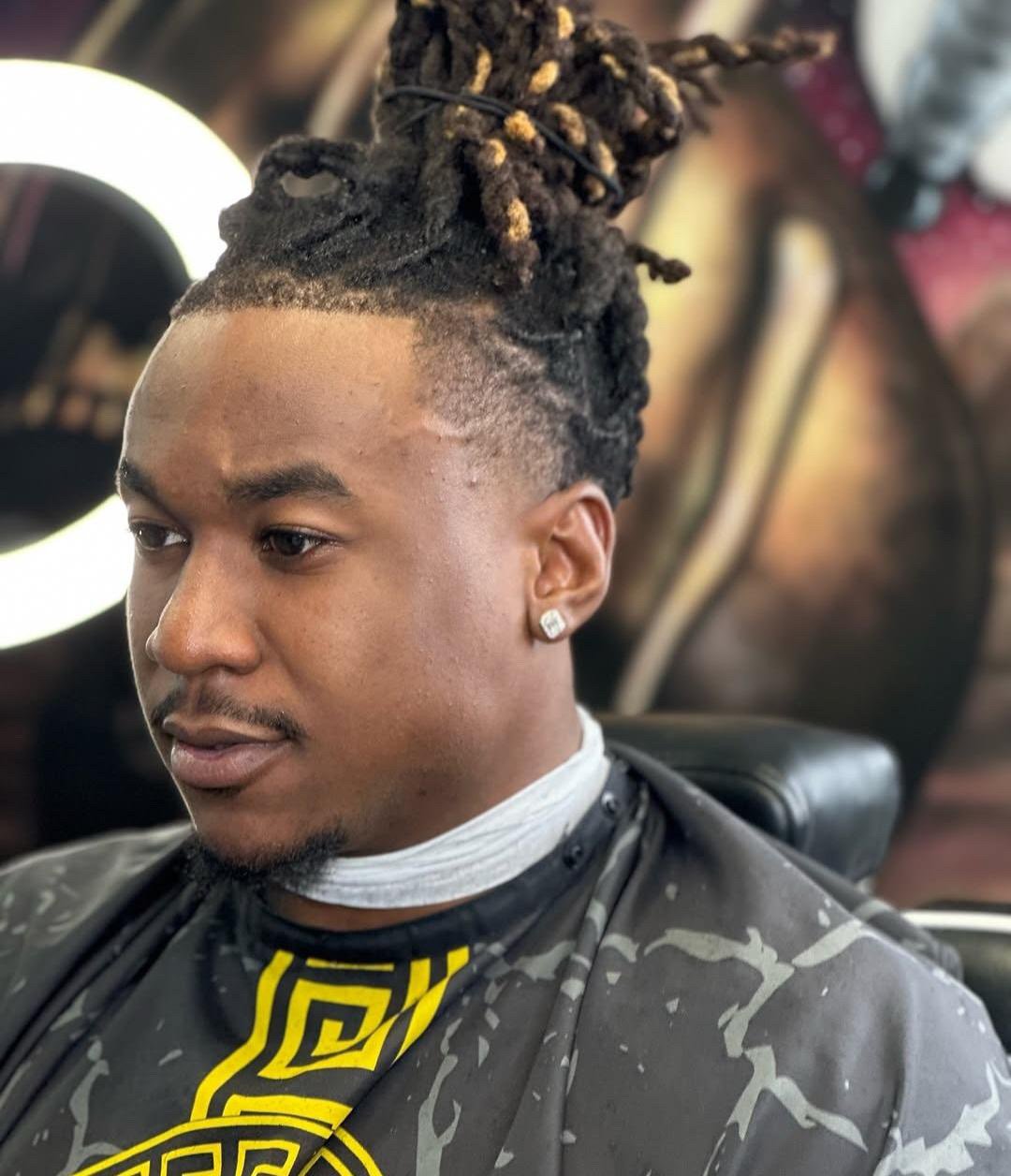 Young man with dreadlocks styled in a top bun, wearing a black and yellow patterned shirt, earrings, and sitting in a barber chair.