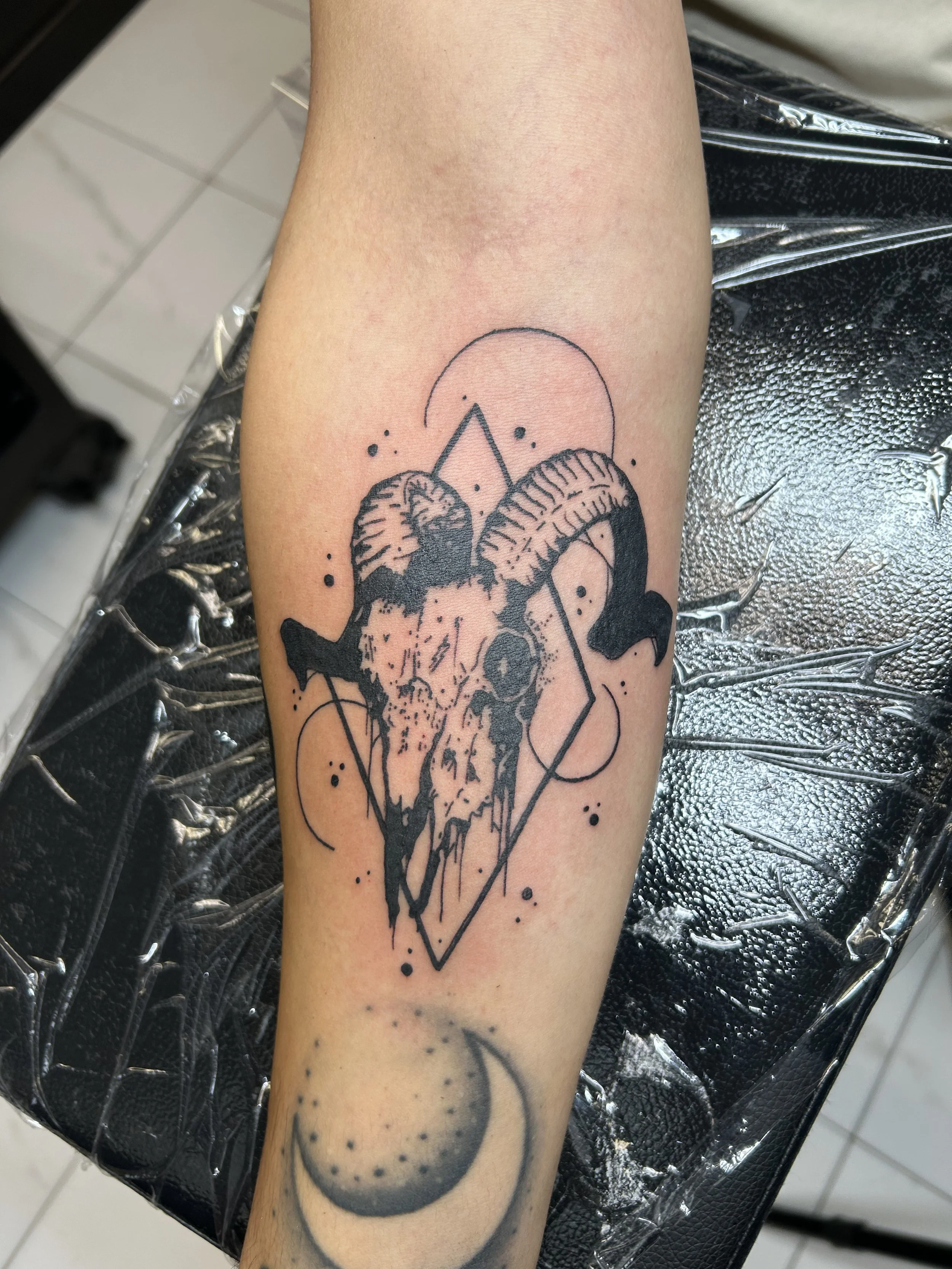 Tattoo of a deer skull with horns, over geometric shapes, circles, and dots, on a person's forearm.