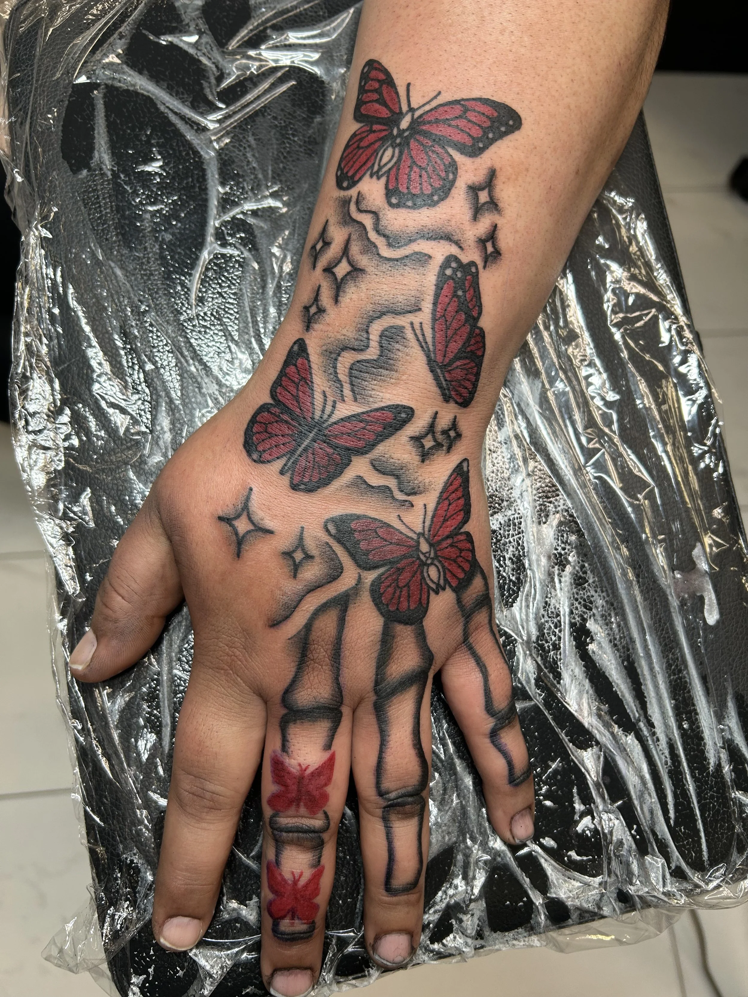 A person’s hand with tattoos of four red butterflies, with black outlines and details, surrounded by black stars and sparkles, and the fingers have black ink tattoos of cracked lines and red butterfly tattoos.