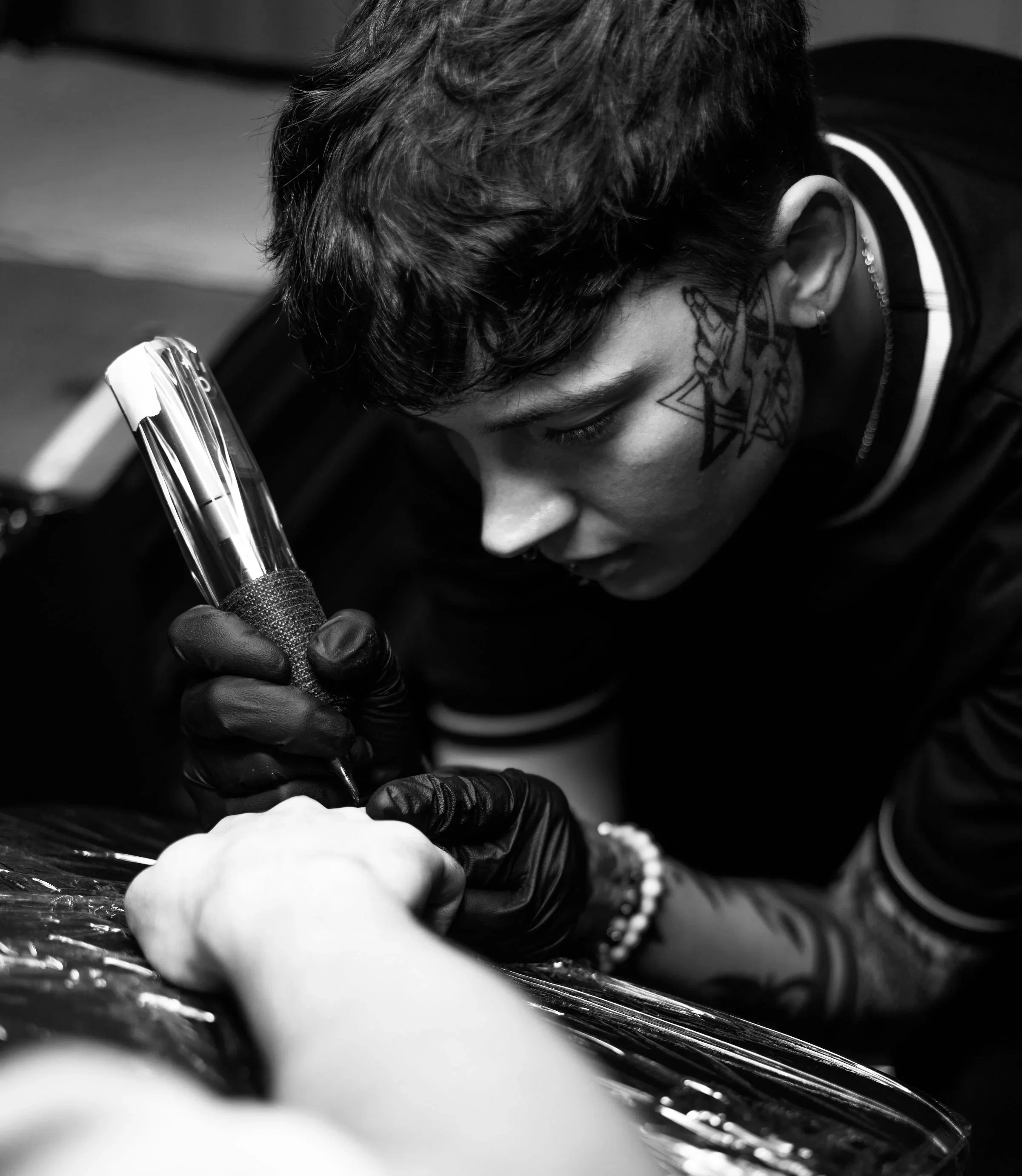 A person with short hair, tattoos, and a nose ring, wearing gloves, is focused on tattooing another person's arm with a tattoo machine.