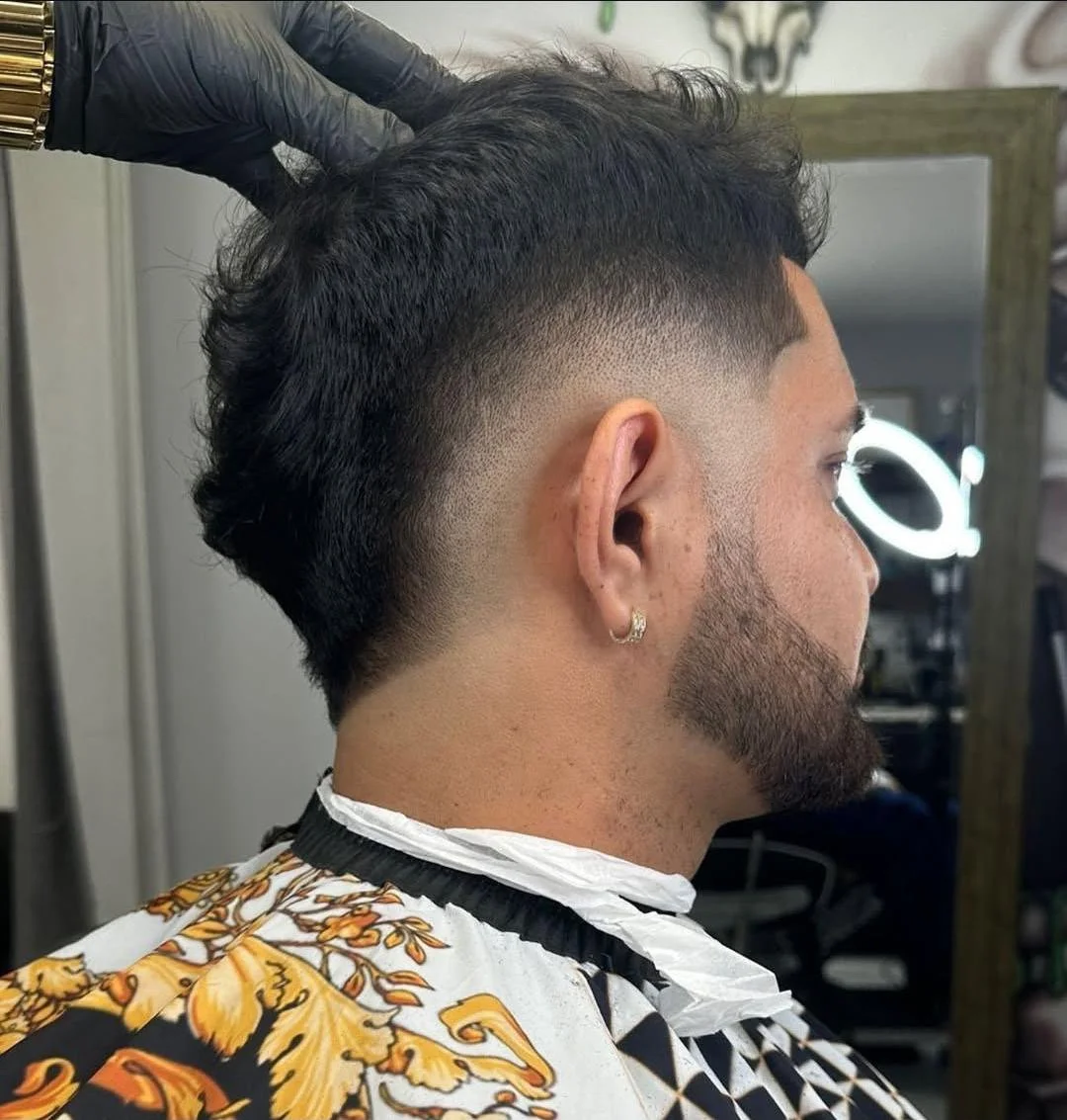 Man getting a fade haircut at a barbershop, wearing a barber cape with a floral pattern, a black glove on one hand, and a ring on his ear.