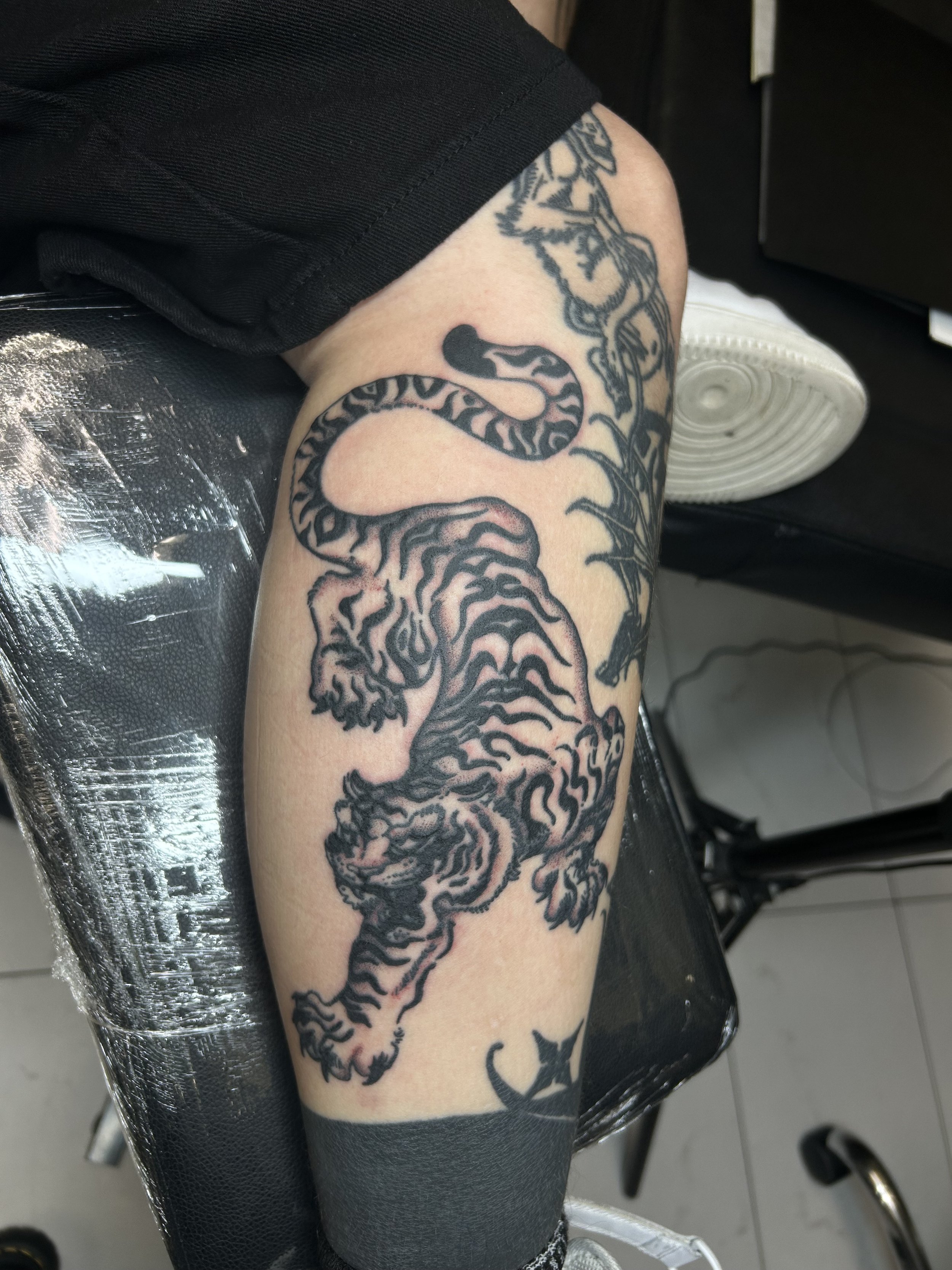 Tattoo of a snake on a person's leg, with leaves and stems around it, sitting on a black chair with a silver surface underneath. The person is wearing black shorts and a white sneaker.