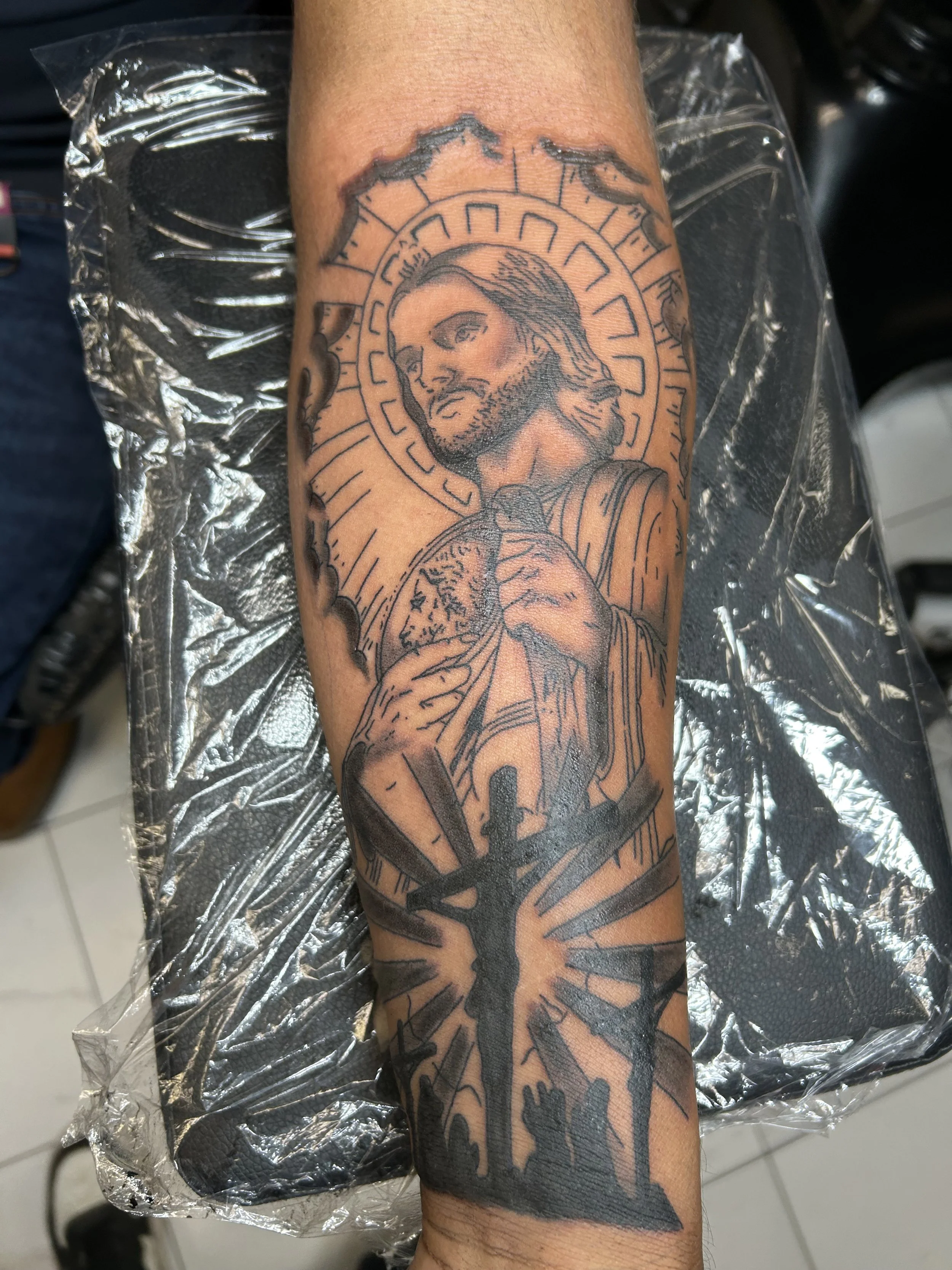 Tattoo of Jesus Christ holding a wound, with a halo and rays around his head, and a crucifix with praying hands beneath him.
