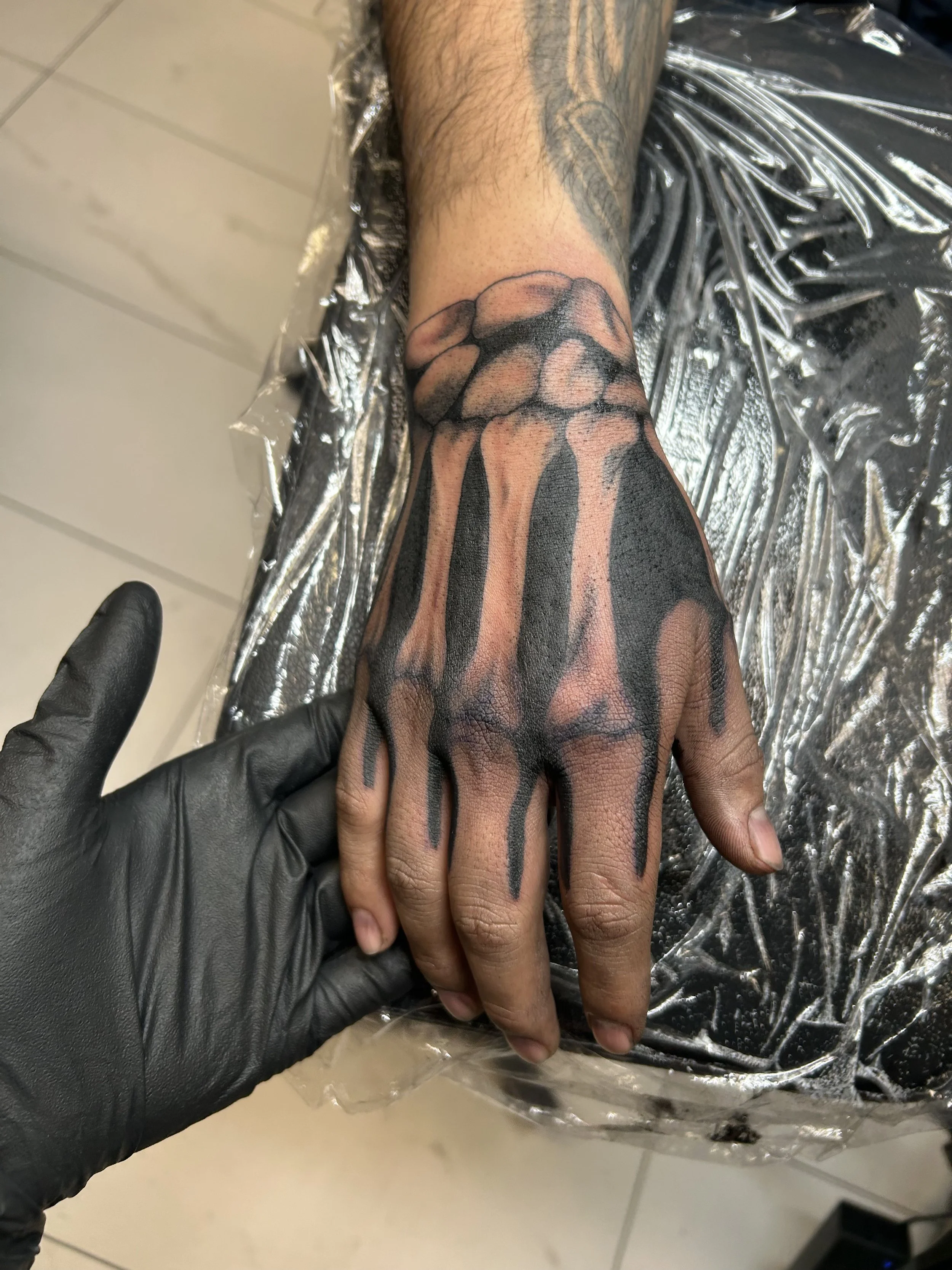 Close-up of a tattoo on a person's hand and wrist, featuring black ink drips coming from a pattern on the hand, with another tattoo partially visible on the forearm.
