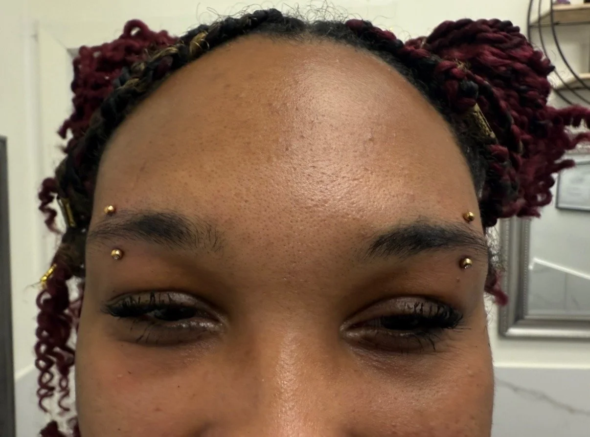 Close-up of a woman's forehead and eyes with decorative facial piercings above her eyebrows, curly red hair styled in twists, and a background of bathroom or clinic setting.