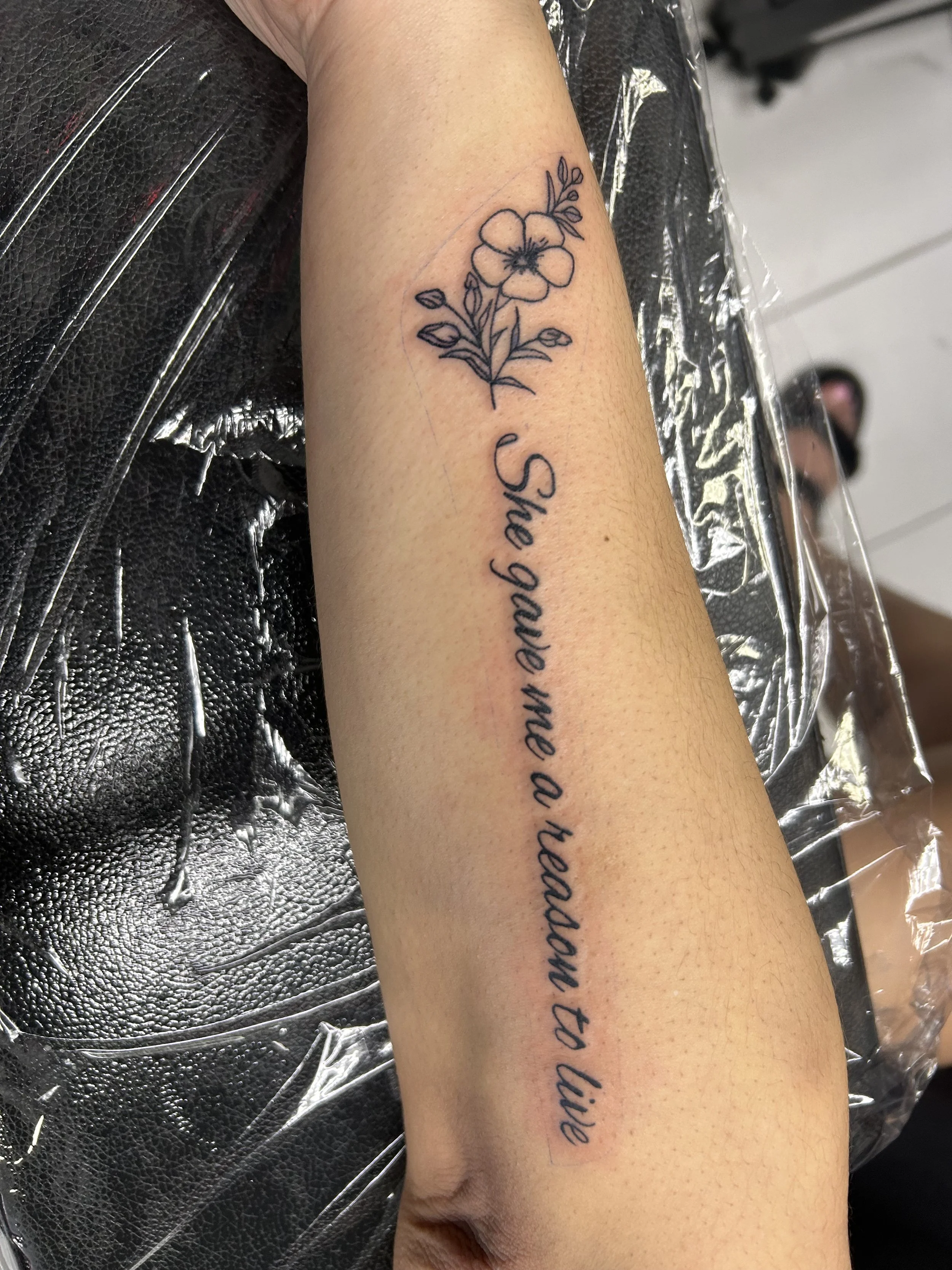 A tattoo on a person's forearm of a flower and the text 'She gave me a reason to live' in cursive script.