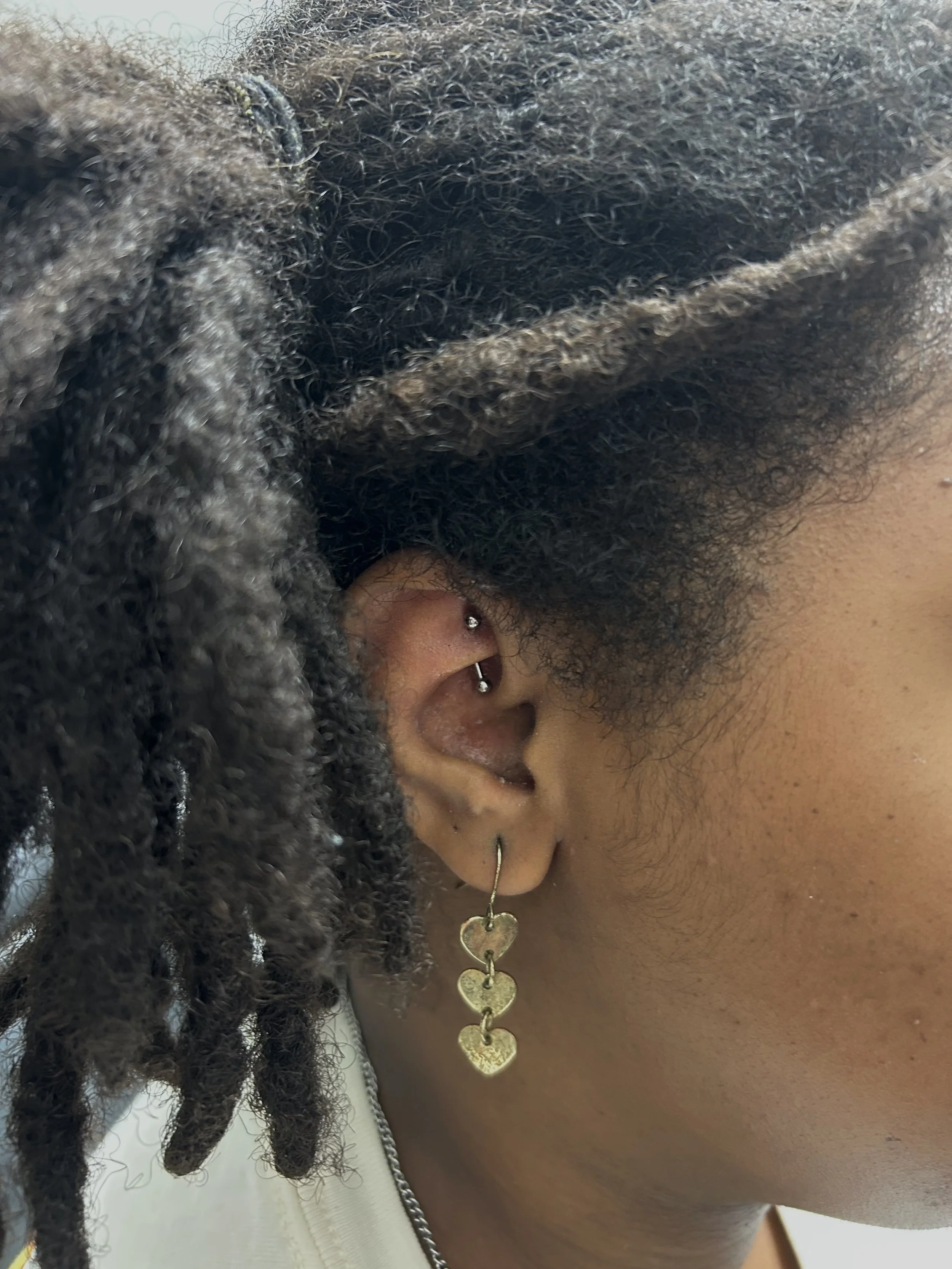 Close-up of a person's ear with two piercings, wearing a black and gray fuzzy hood, with curly black hair and gold heart-shaped jewelry on their necklace.