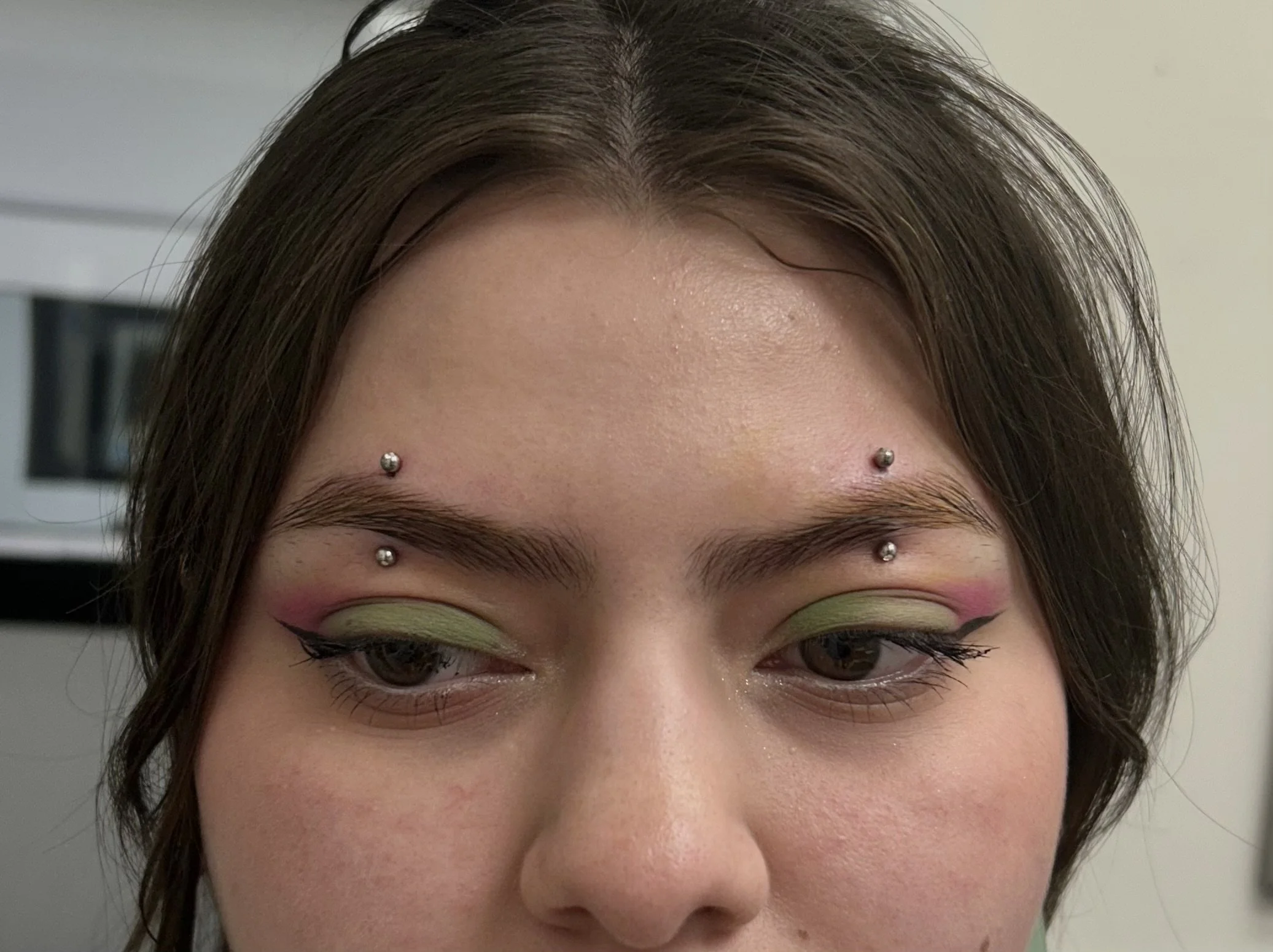 Close-up of a woman's forehead with two dermal piercings, with colorful eye makeup and styled hair.