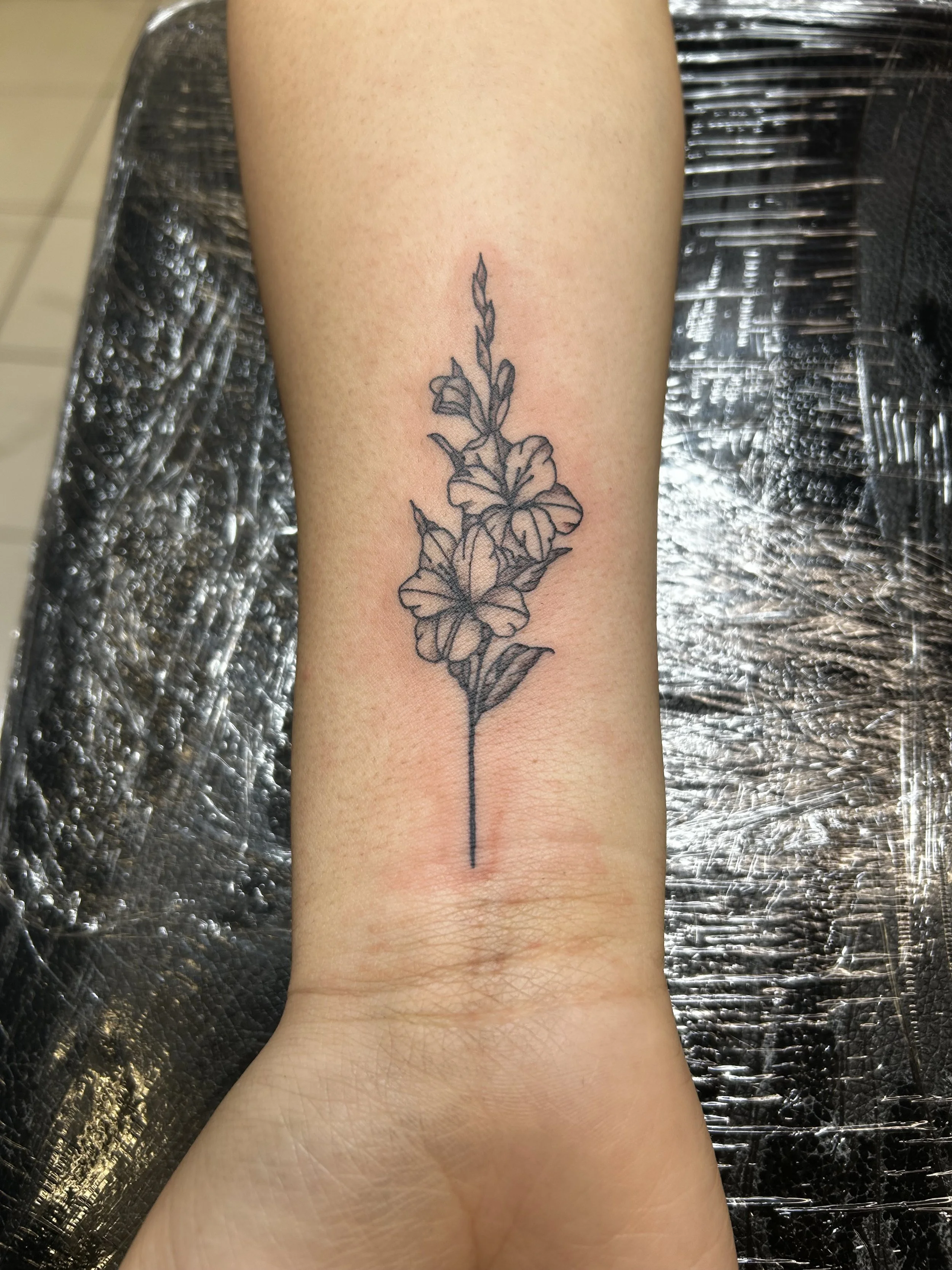 A black ink line tattoo of a floral design on a person's forearm, featuring a vertical stem with leaves and blossoms.