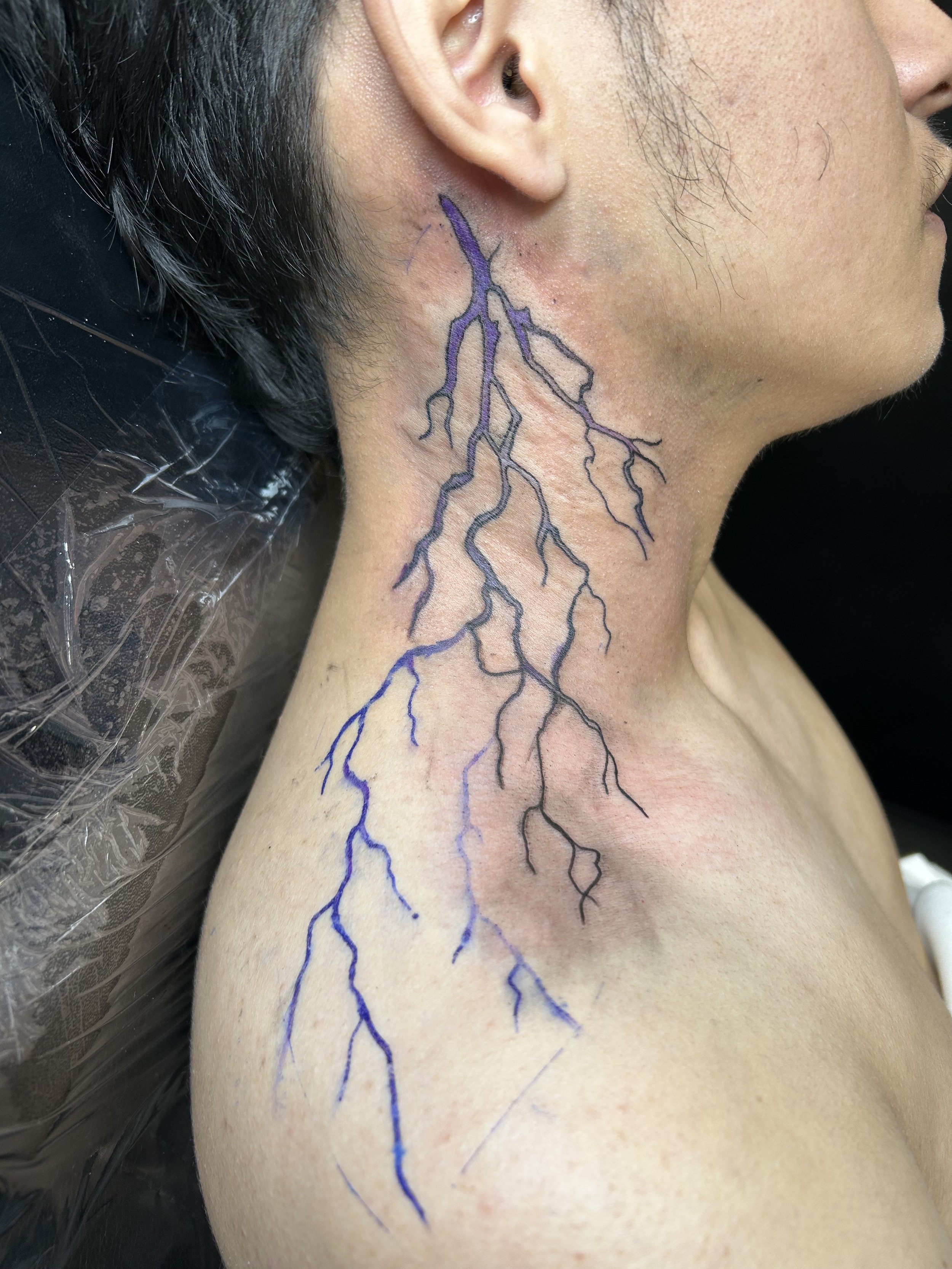 A person has a tattoo of a blue and black lightning bolt design running from the neck down onto the shoulder.