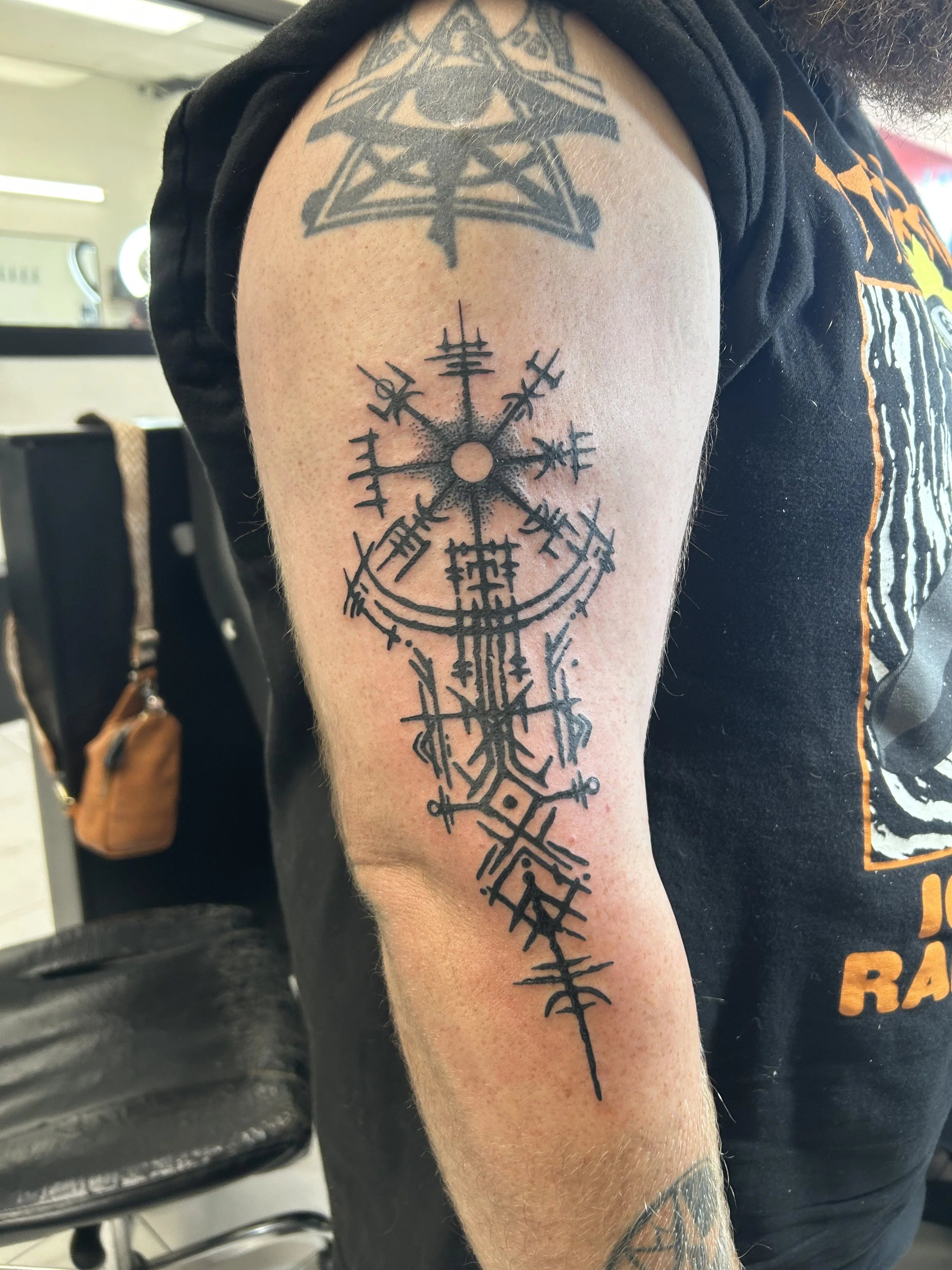 Tattoo of a Vegvisir, a Nordic symbol, on a person's upper arm with other Nordic symbols above and below it.