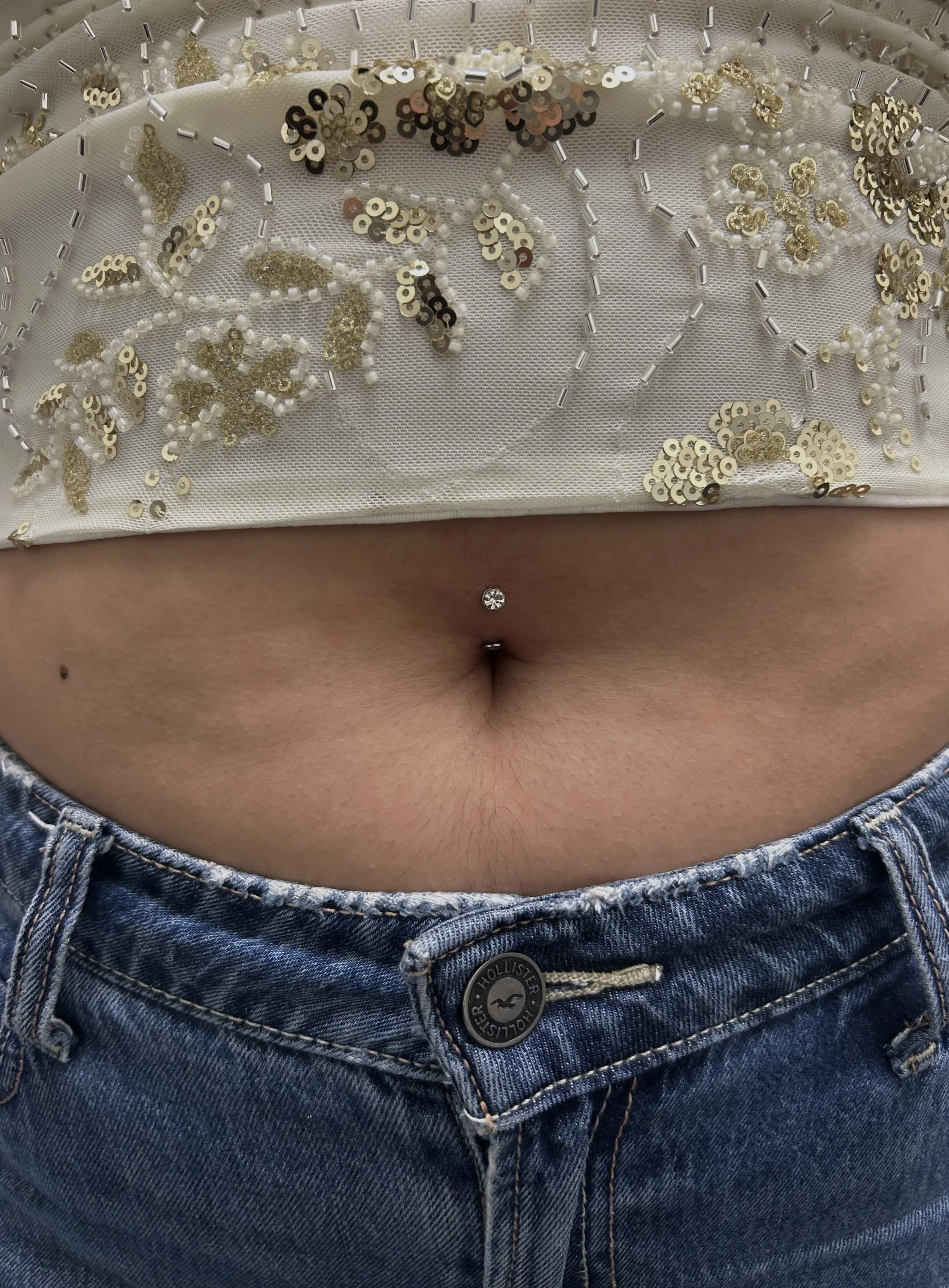 Close-up of a person's midsection wearing distressed blue jeans and a white, sequined, beaded top with floral designs. The person's belly button is pierced with a small, clear gemstone.