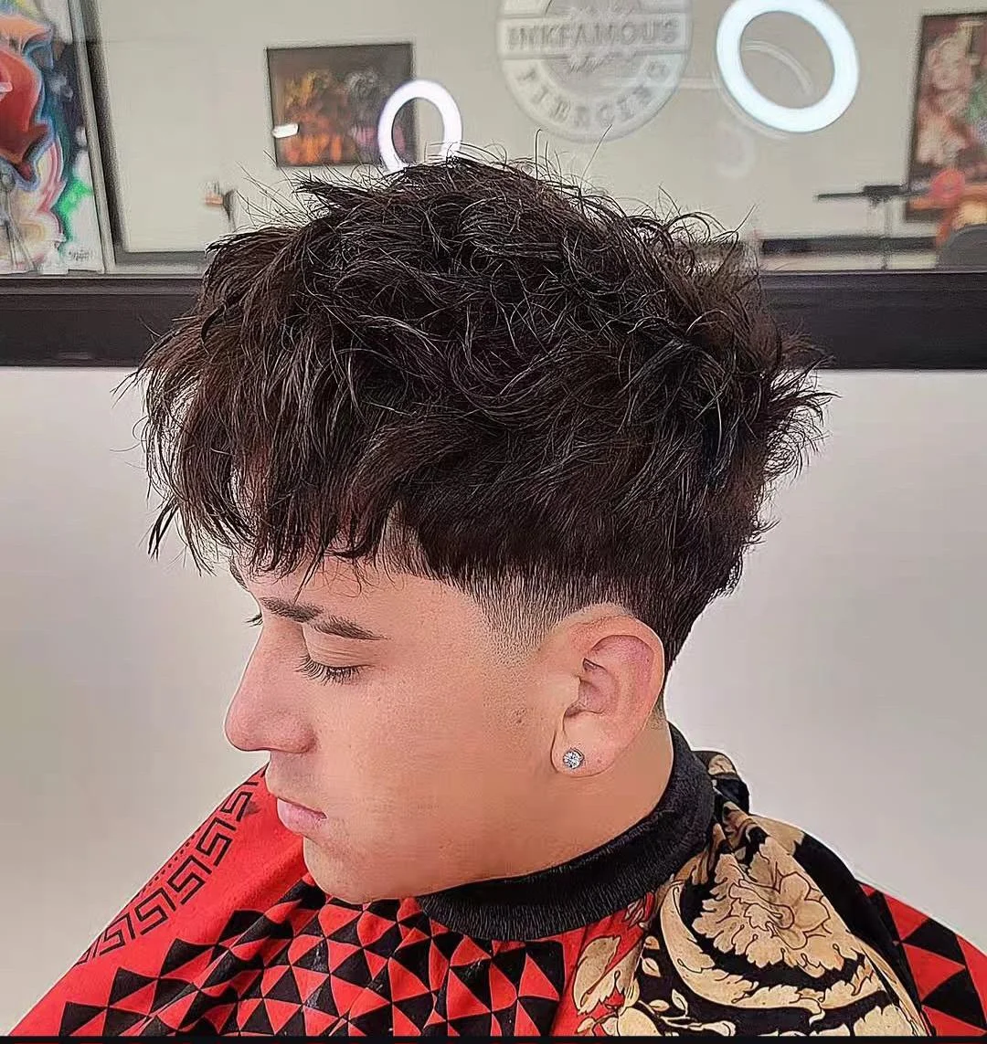 Young man with a short fade haircut and curly hair on top, wearing a black and gold patterned cape, sitting with eyes closed in a hair salon.