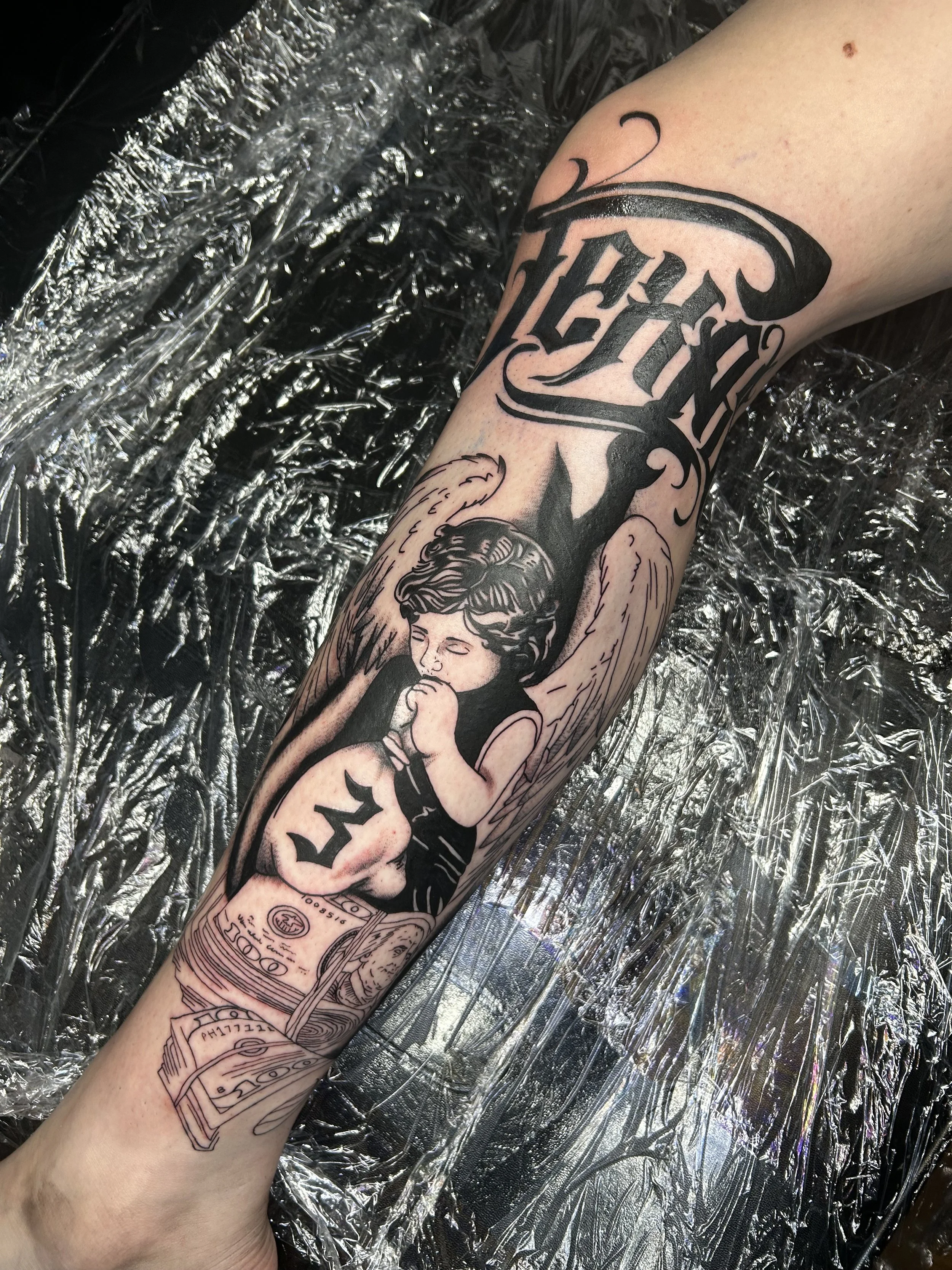 A person's forearm tattoo featuring a woman with angel wings, surrounded by stylized text, with a background of a tattoo studio surface covered in metallic foil.