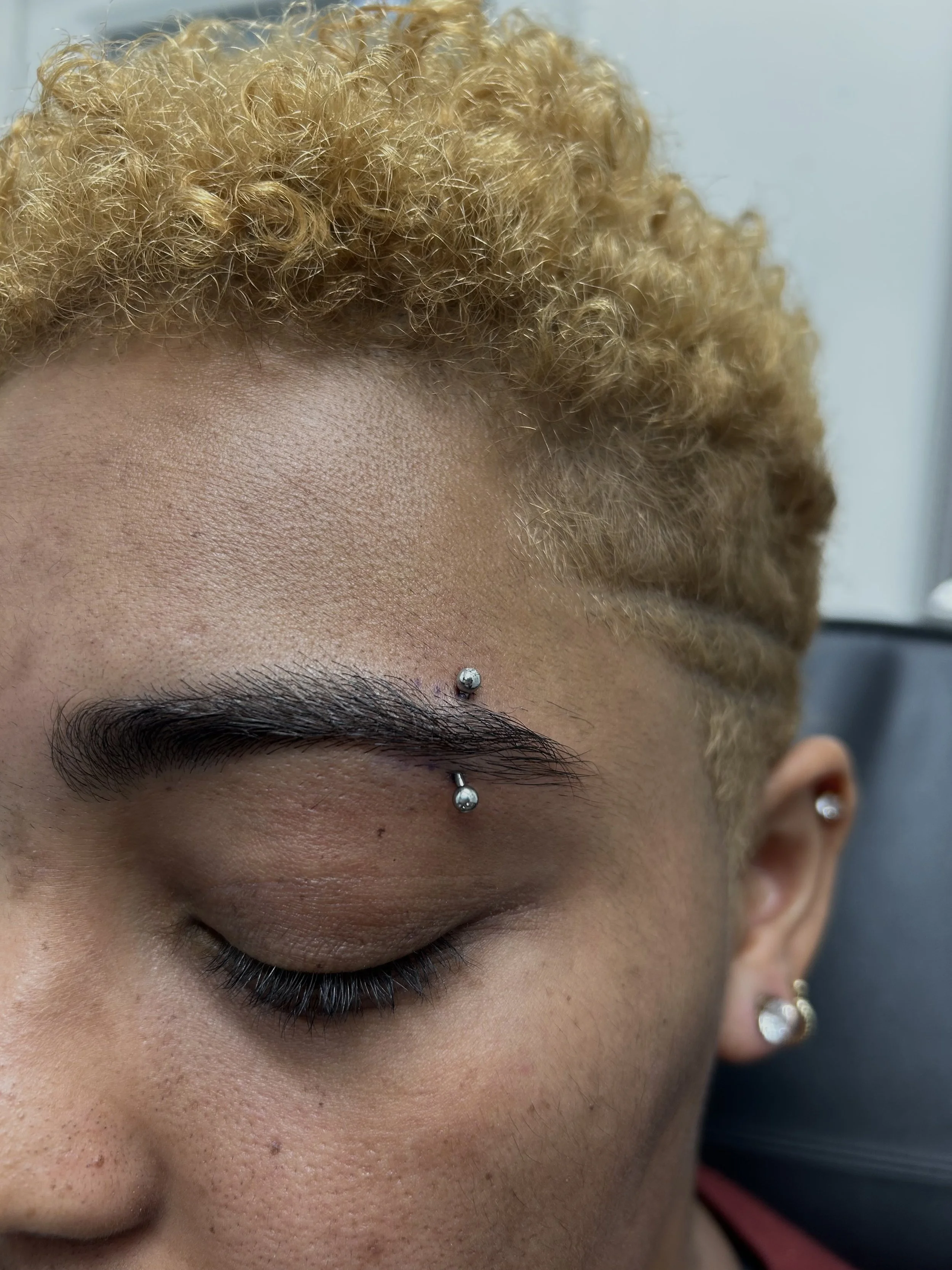 Close-up of a person's face with blond curly hair, a well-groomed eyebrow, and multiple facial piercings including a nose stud, a vertical eyebrow bar, and earrings.