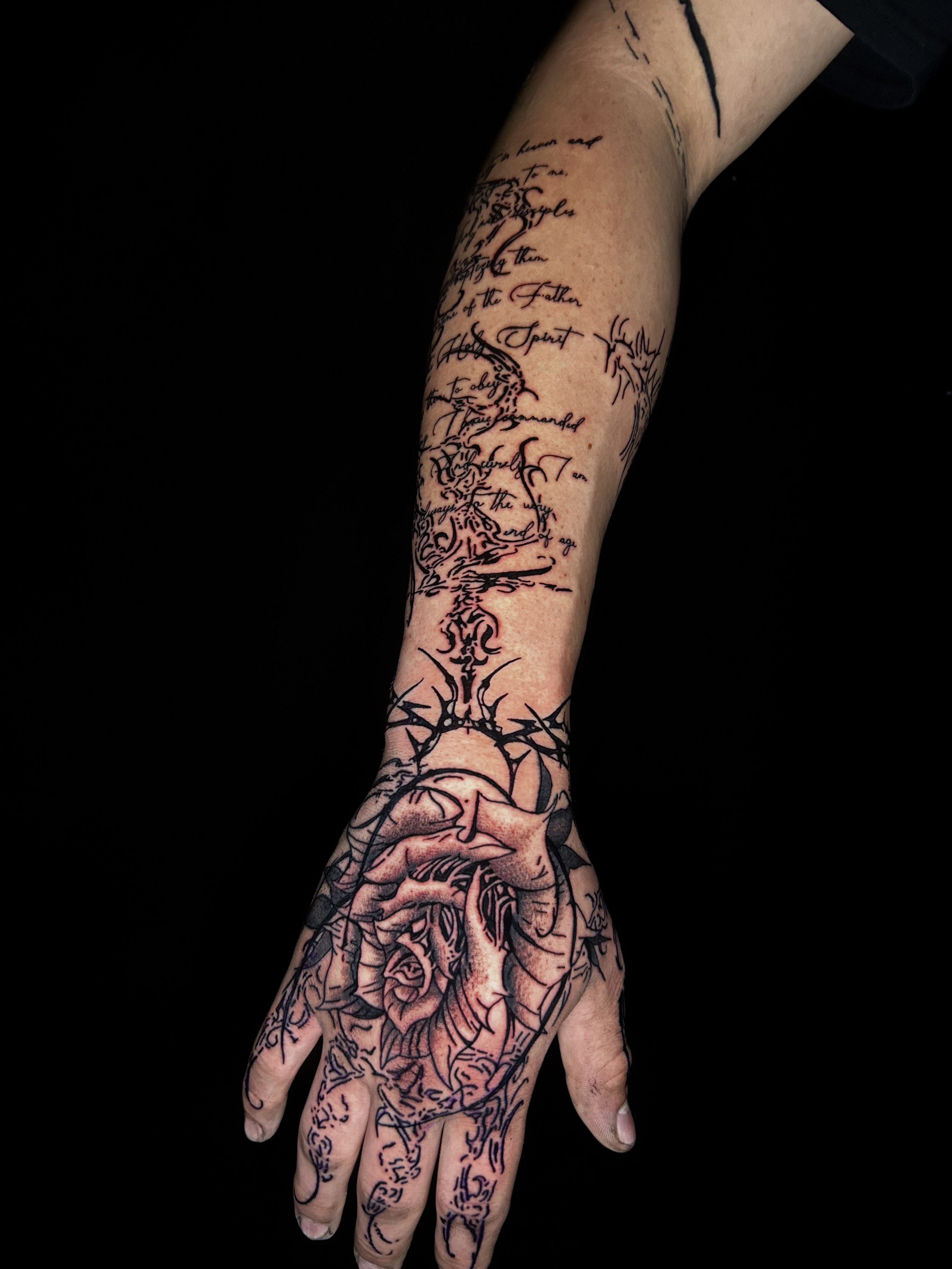 Tattooed arm and hand with a detailed heart design and handwritten script on the forearm, against a black background.