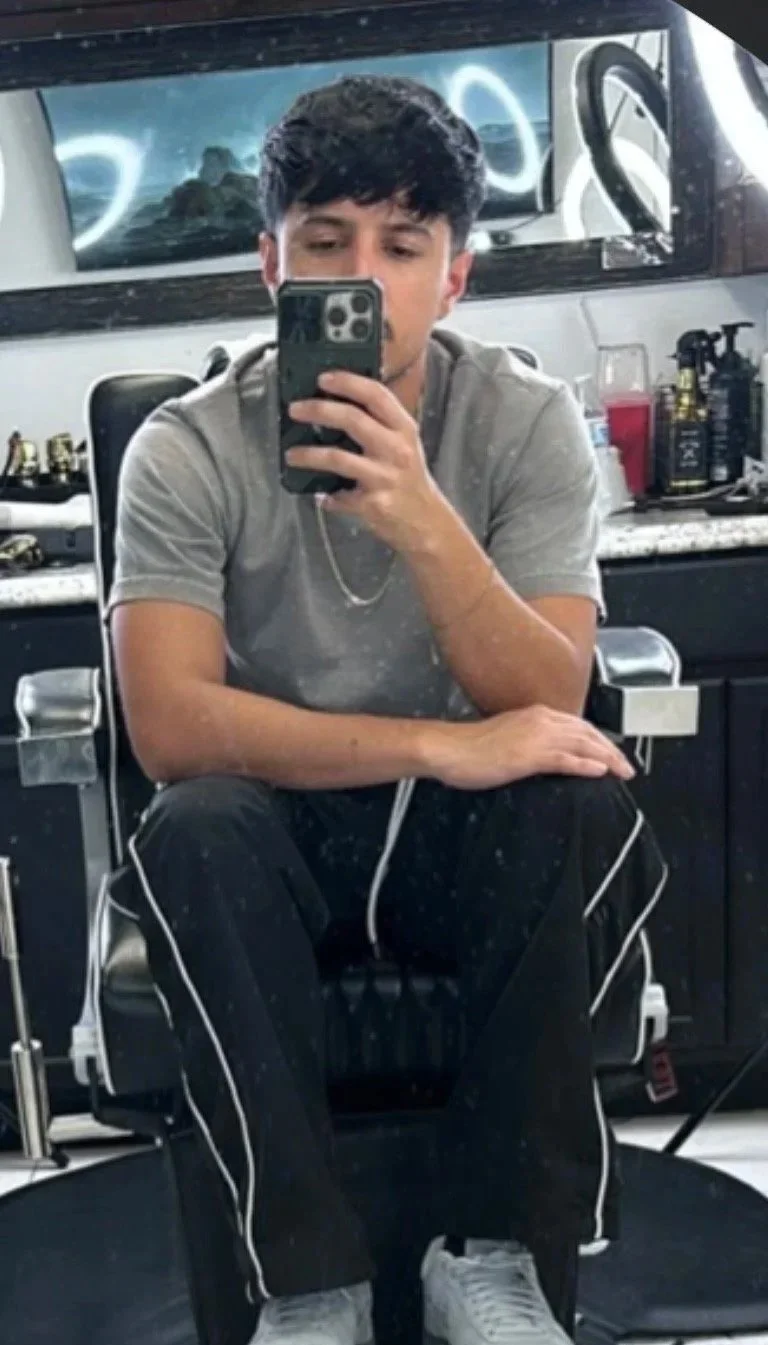 A young man taking a selfie in a barber shop mirror, sitting on a chair with a black and gray interior, hair products on the counter behind him, wearing a gray T-shirt, black pants with white piping, and white sneakers.