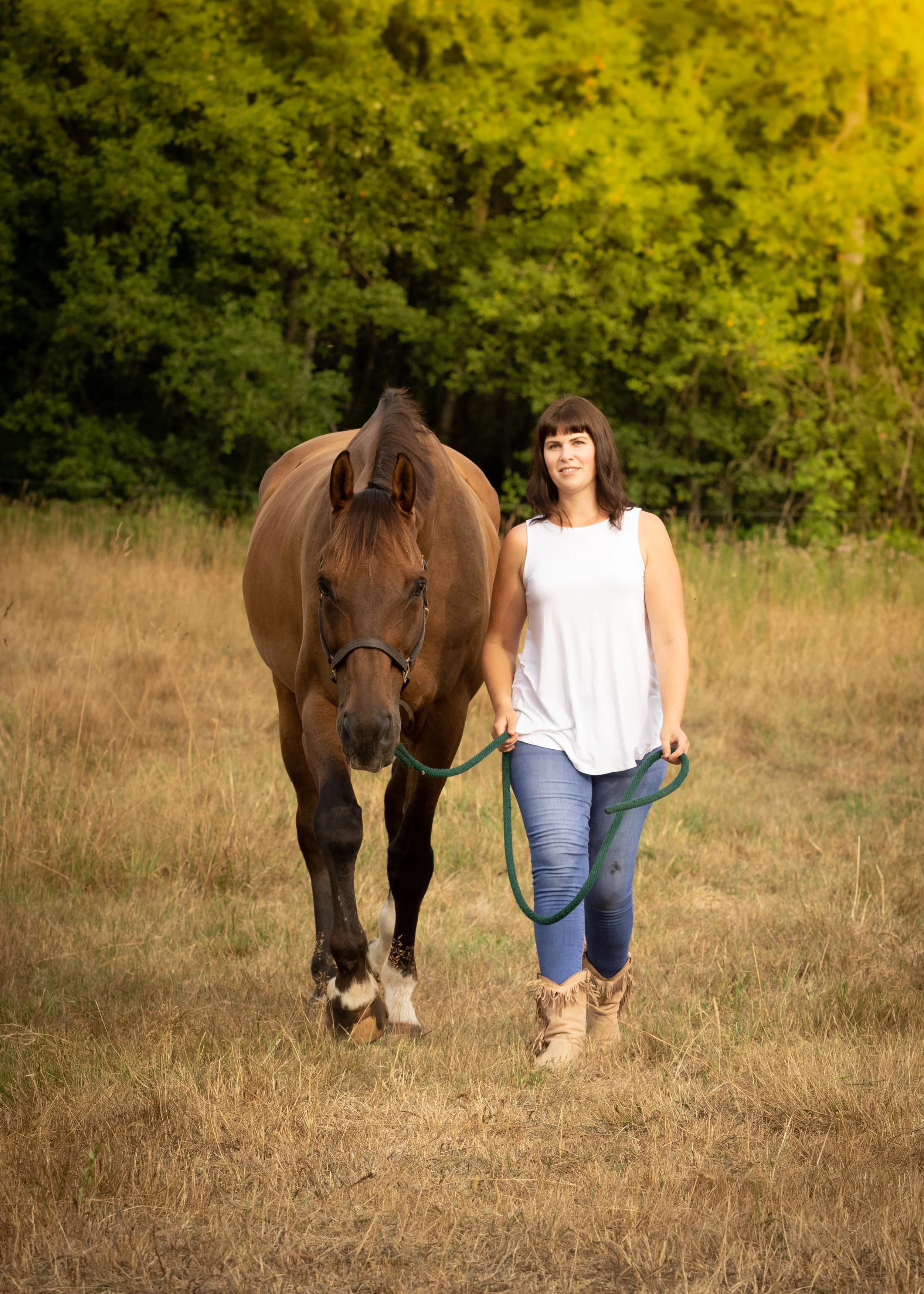 A woman walking with a brown horse in a grassy field with green trees in the background.