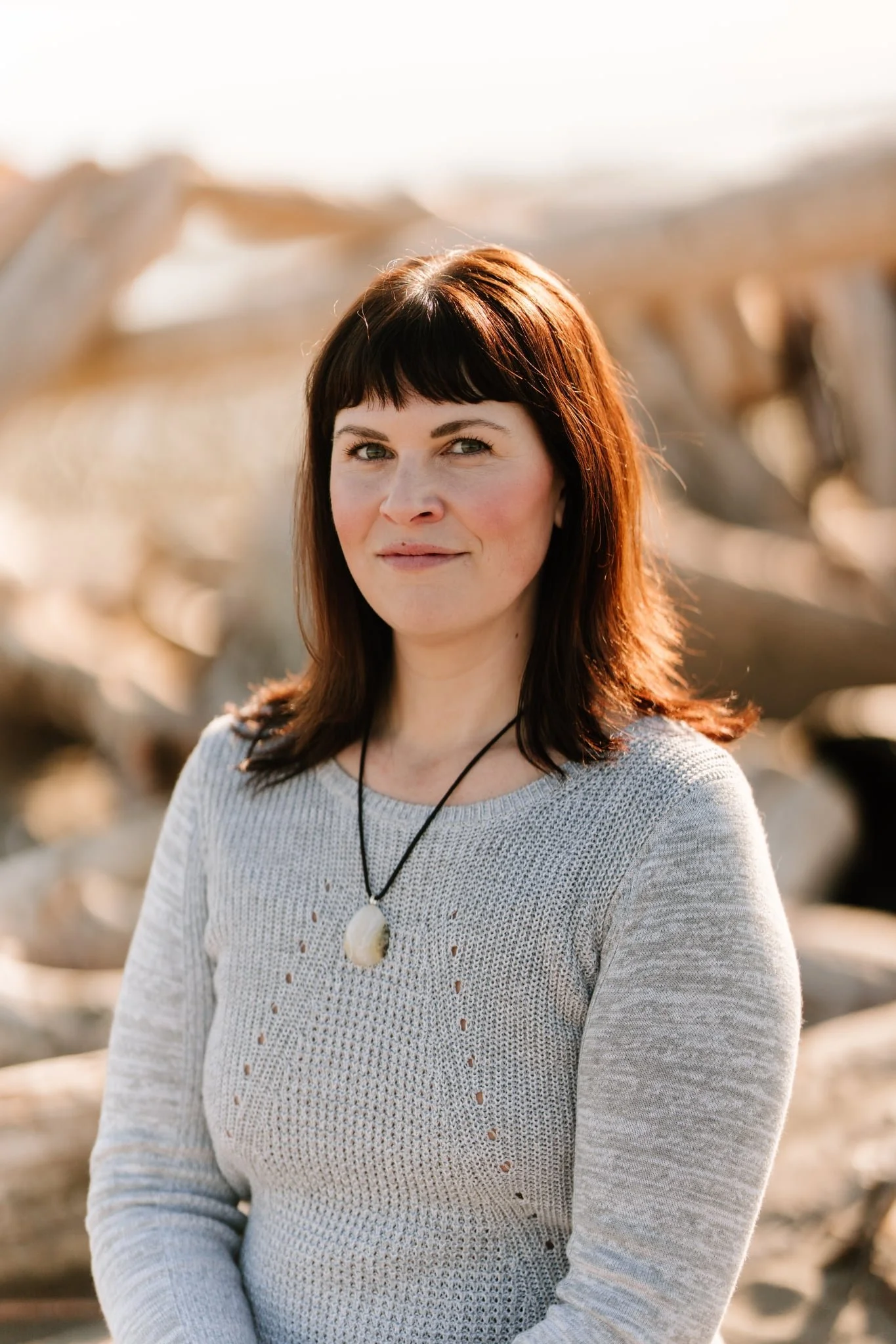 A woman with medium-length brown hair and bangs standing outdoors in warm sunlight, wearing a light-colored textured sweater and a necklace with a large pendant, with blurred rocks or driftwood in the background.