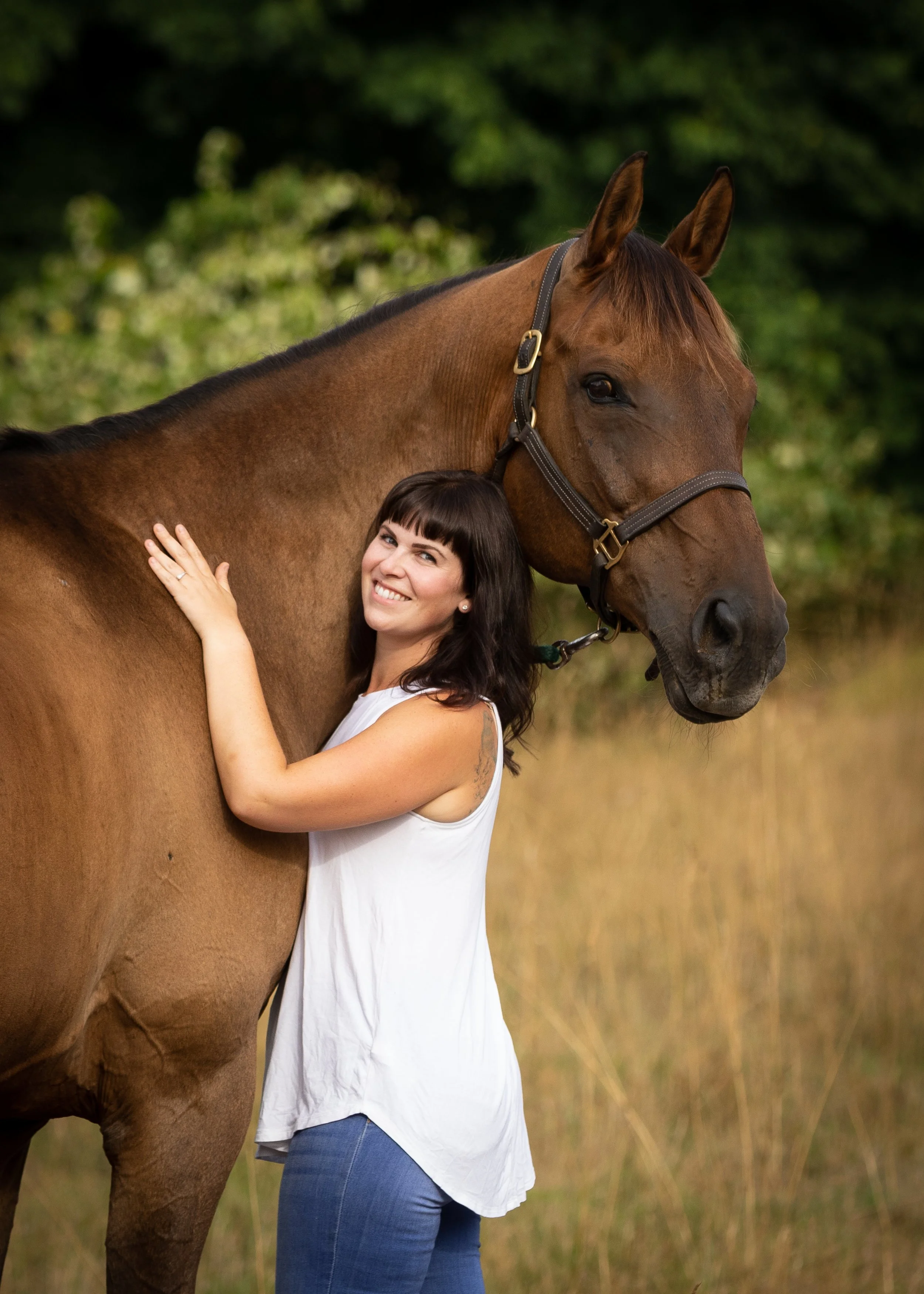 A woman with dark hair, wearing a white sleeveless top and blue jeans, hugging a brown horse with a black mane outdoors, with green trees in the background.