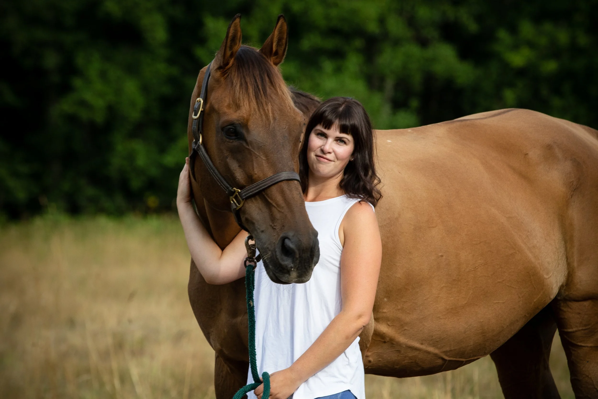 A woman with brown hair wearing a white tank top hugging a brown horse outdoors with a green blurred background.
